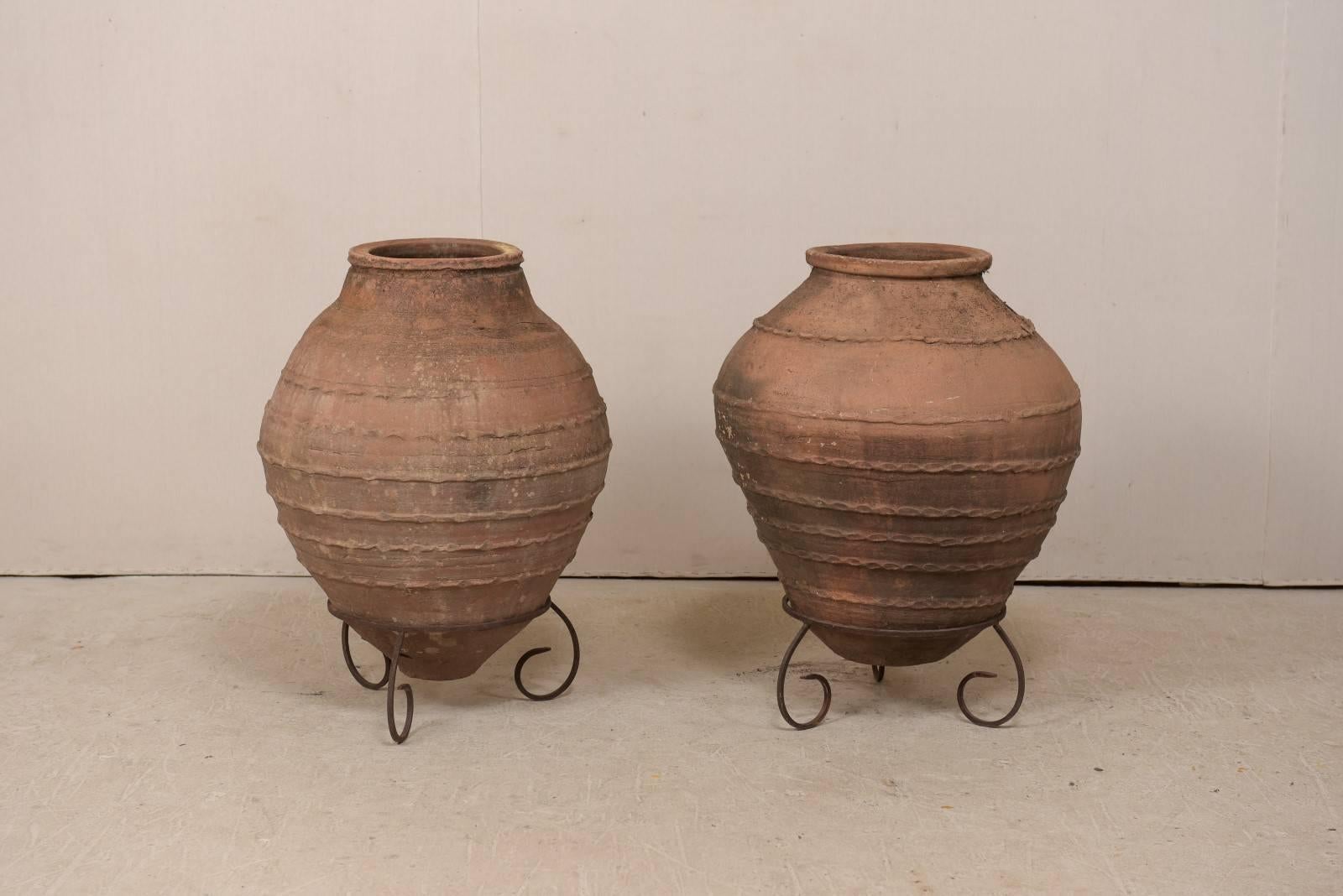 A pair of antique Turkish olive jars on custom stands. This pair of antique Turkish vessels, from the early 20th century, were once used to hold olives. These rounded terracotta vessels have bulbous centers which taper to their bottoms, and are