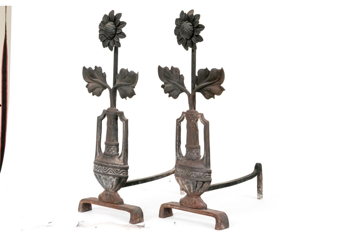 Pair of wrought iron andirons in Arts and Crafts style presenting in the form of sunflowers emitting from a handled urn on a plinth base.

Dimensions: 10 1/8