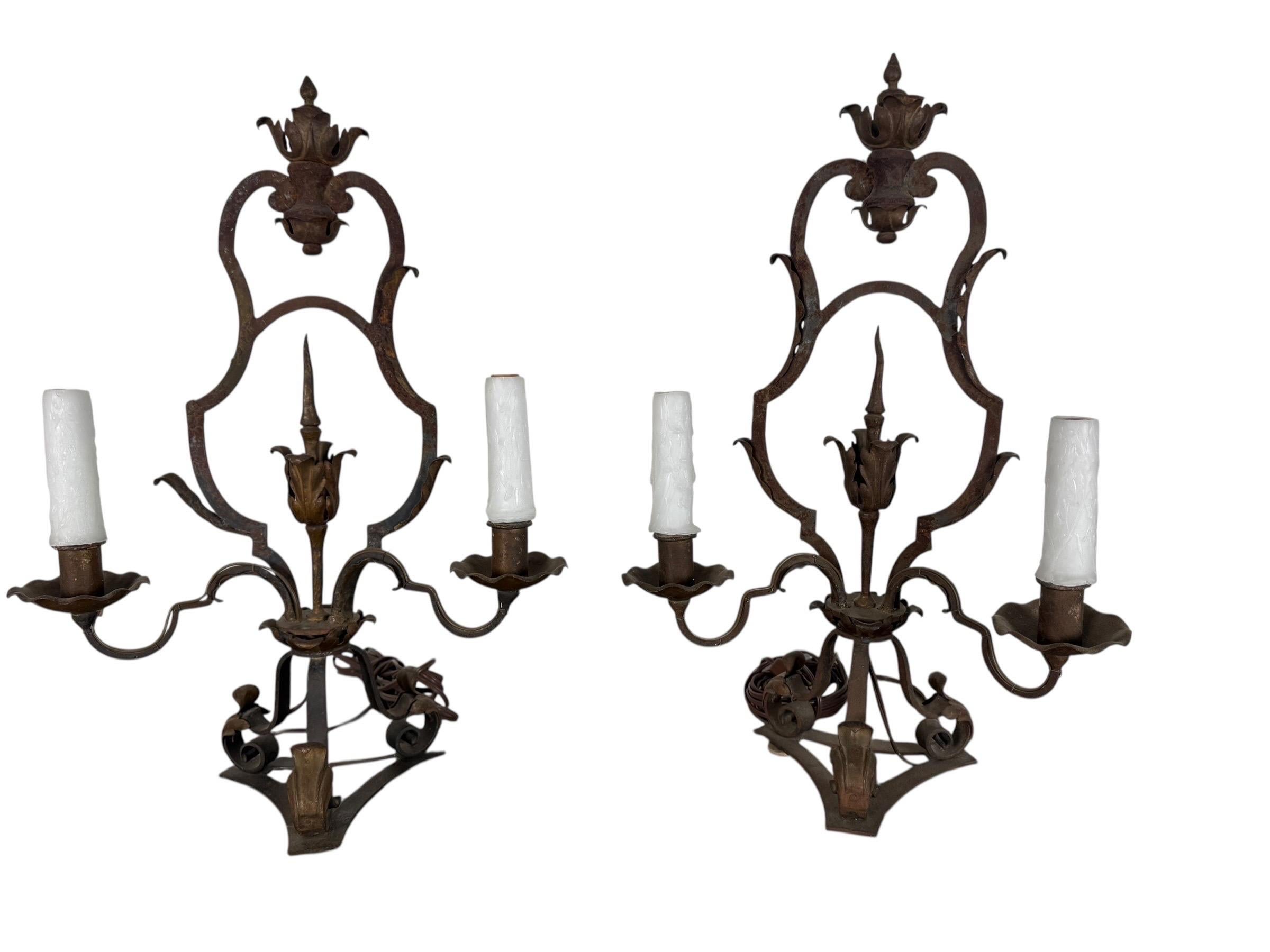 A striking pair of hand-forged wrought iron lamps in the Spanish Revival taste, dating to the early 20th century. Each sconce features an elongated shield-form back crowned with a dramatic finial shaped as a stylized budding flame, flanked by
