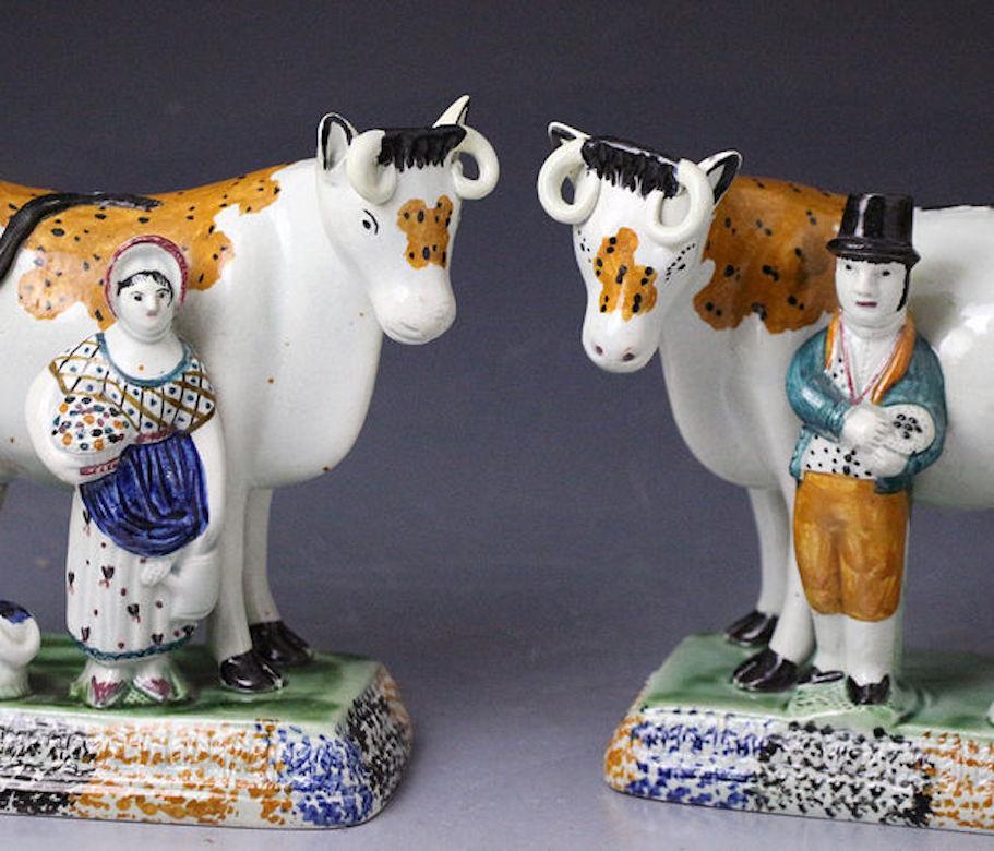 A pair of Pratt colored figures of standing cows with male and female attendants, calf and dog modeled on an oblong stepped shape base with speckled orange, black and blue banded border. The cows are decorated with blotches of ochre which are