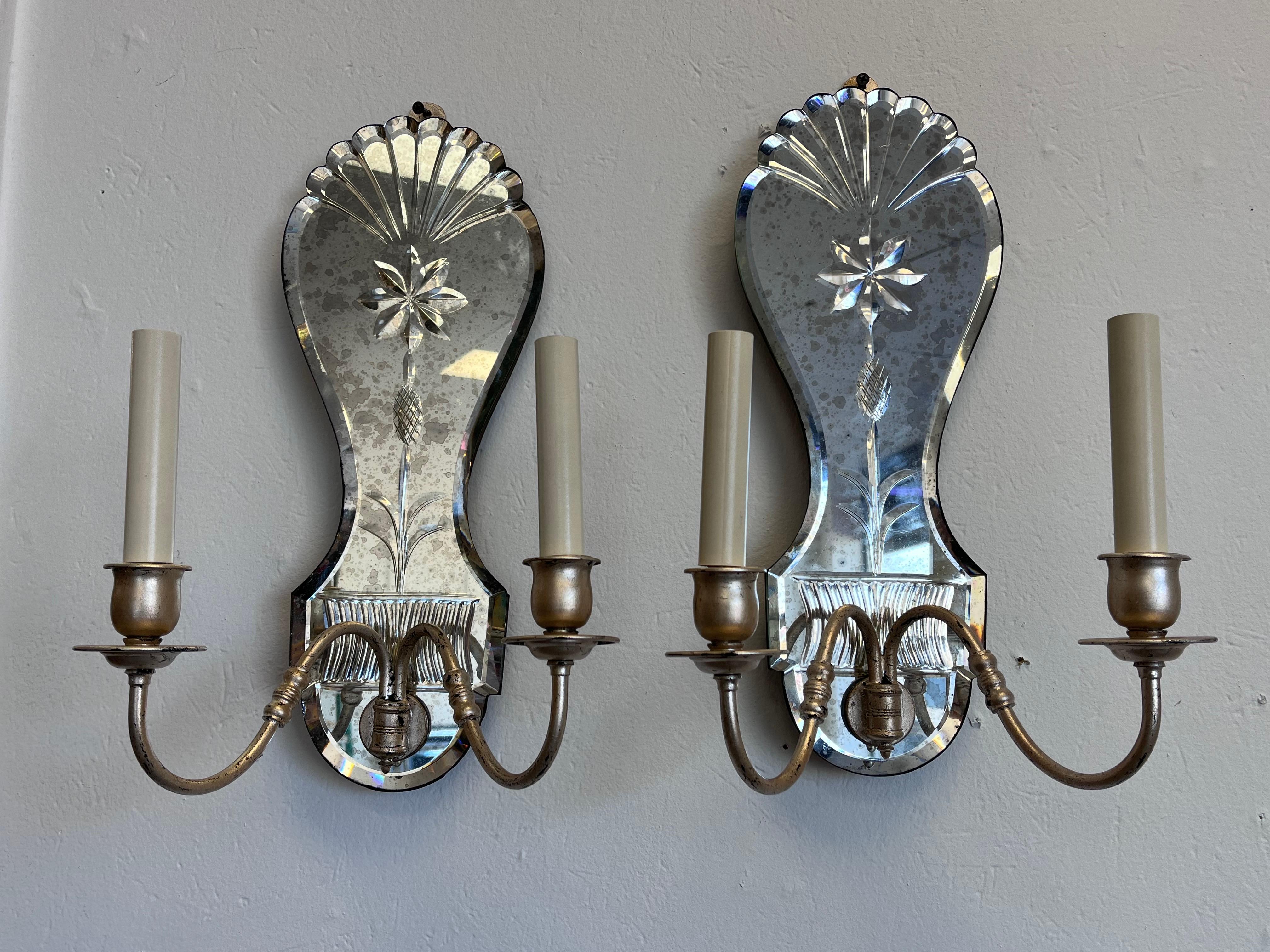 A large pair of ornately designed antiqued and cut mirror wall sconces with silver gilt scrolling, double arms. The top of each of the wall lights is cut in a fan shaped or scalloped design. This element is atop an eight petal flower decor. Moving