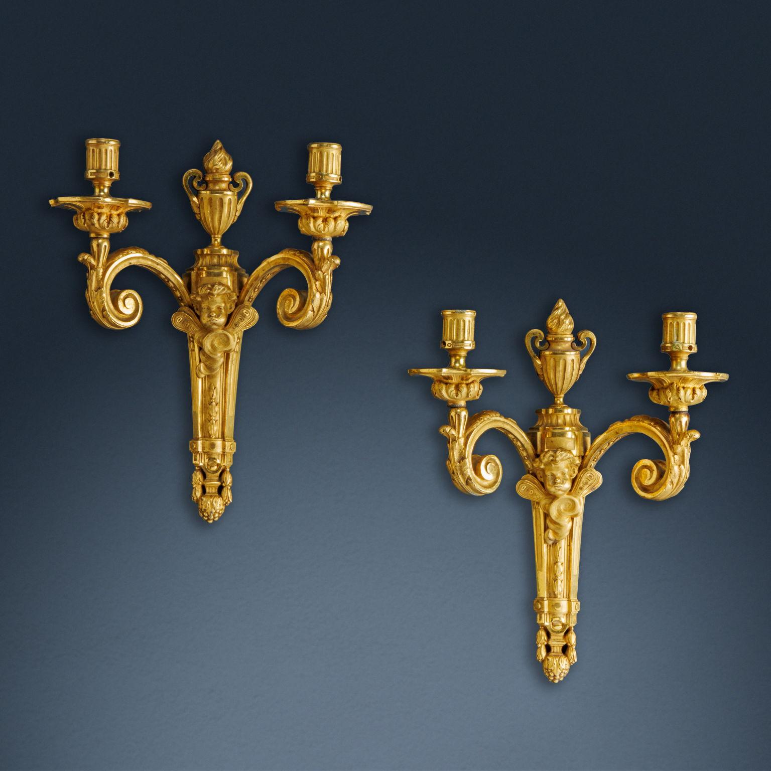 Pair of two-light wall sconces made of chased and gilded bronze. Two arms with leaf volutes take their shape from a central shaft, composed of a tapered semi-column, on which geometric elements alternate with phytomorphic motifs; the pilaster is