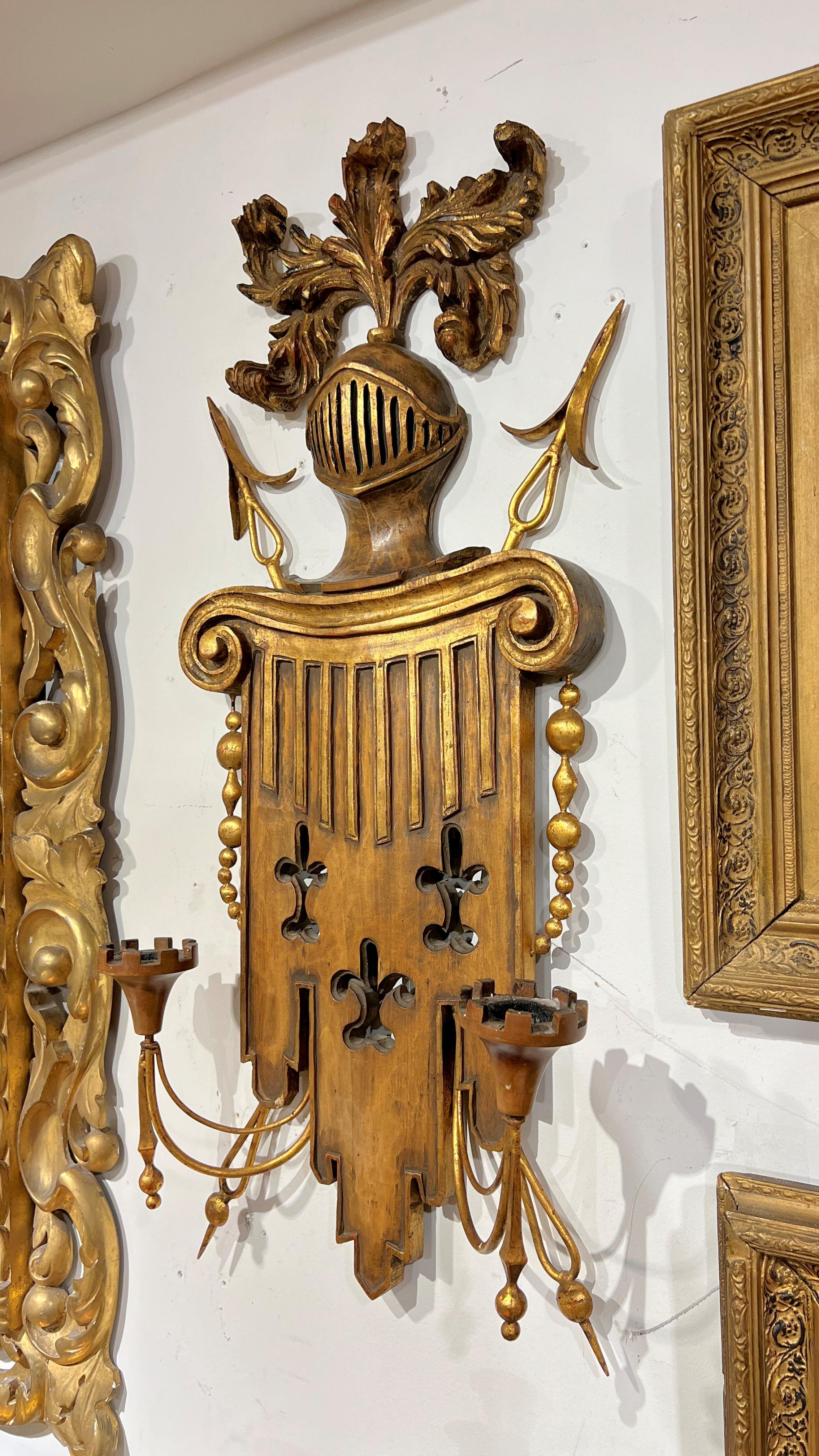 Zwei geschnitzte und vergoldete Vintage-Wandleuchter aus Metall im gotischen Stil mit kunstvoll geschnitzten Rückseiten in Form einer Säule mit Fleur-de-Lis und dorischem Kapitell, darüber ein Ritterhelm mit Federfedern, Schwertspitzen und zwei