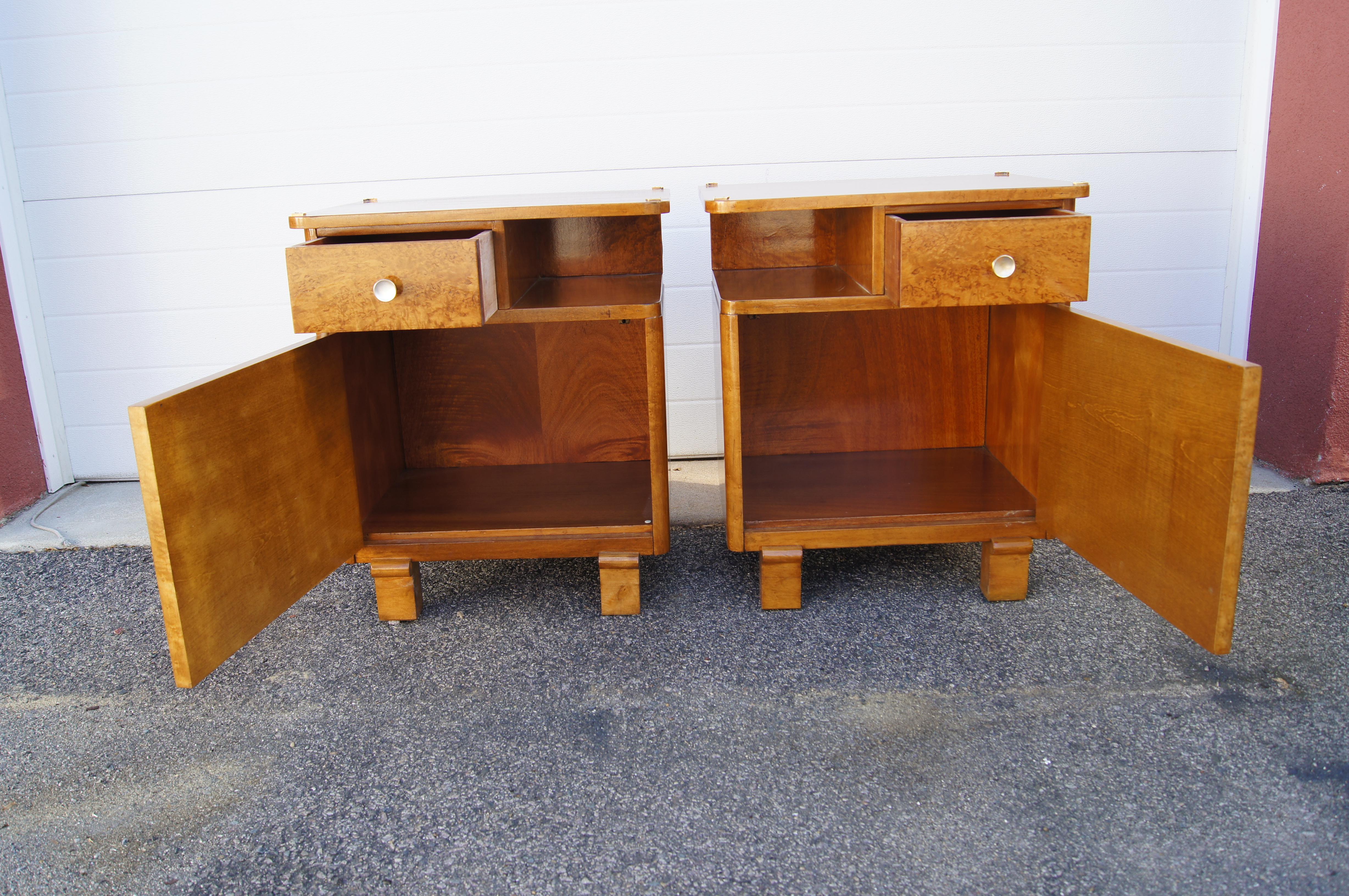 This lovely pair of 1930s Art Deco nightstands features mirror-image glossy burl wood cases on sturdy carved legs. Behind a door is a large storage space; above is a small drawer opposite an open shelf. Brass accents decorate each corner of the top