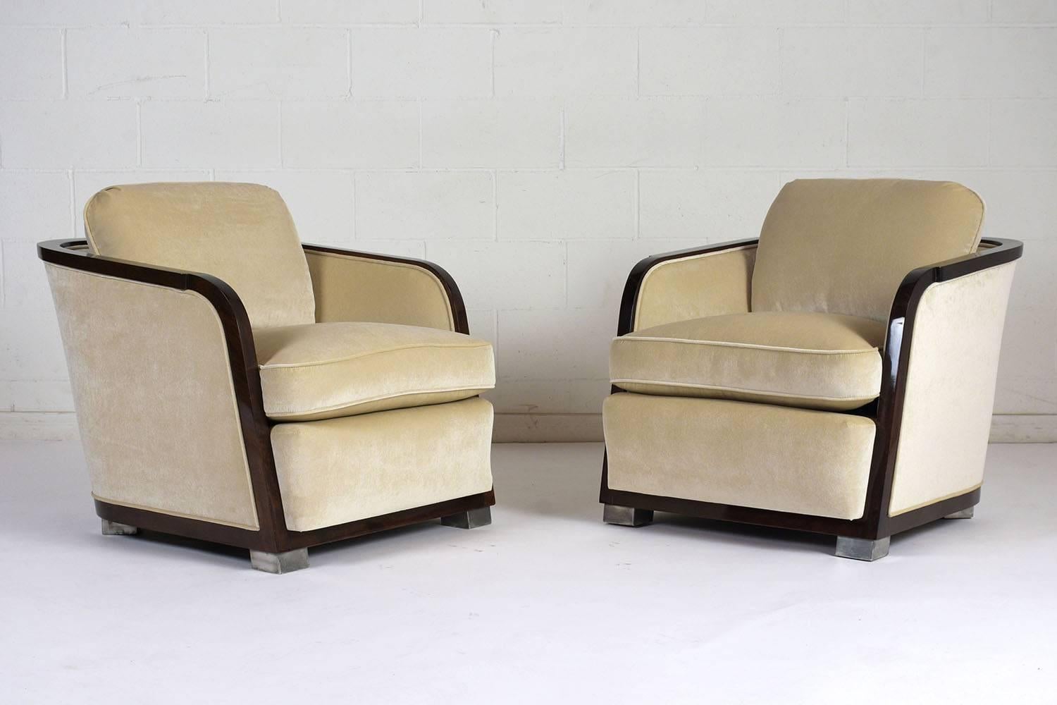 This pair of 1960s Art Deco-style club chairs have a curved profile with a rosewood color wood frame with a lacquered finish. The chairs are upholstered on the back, seat, and front edge in a beige color velvet fabric with single piping trim