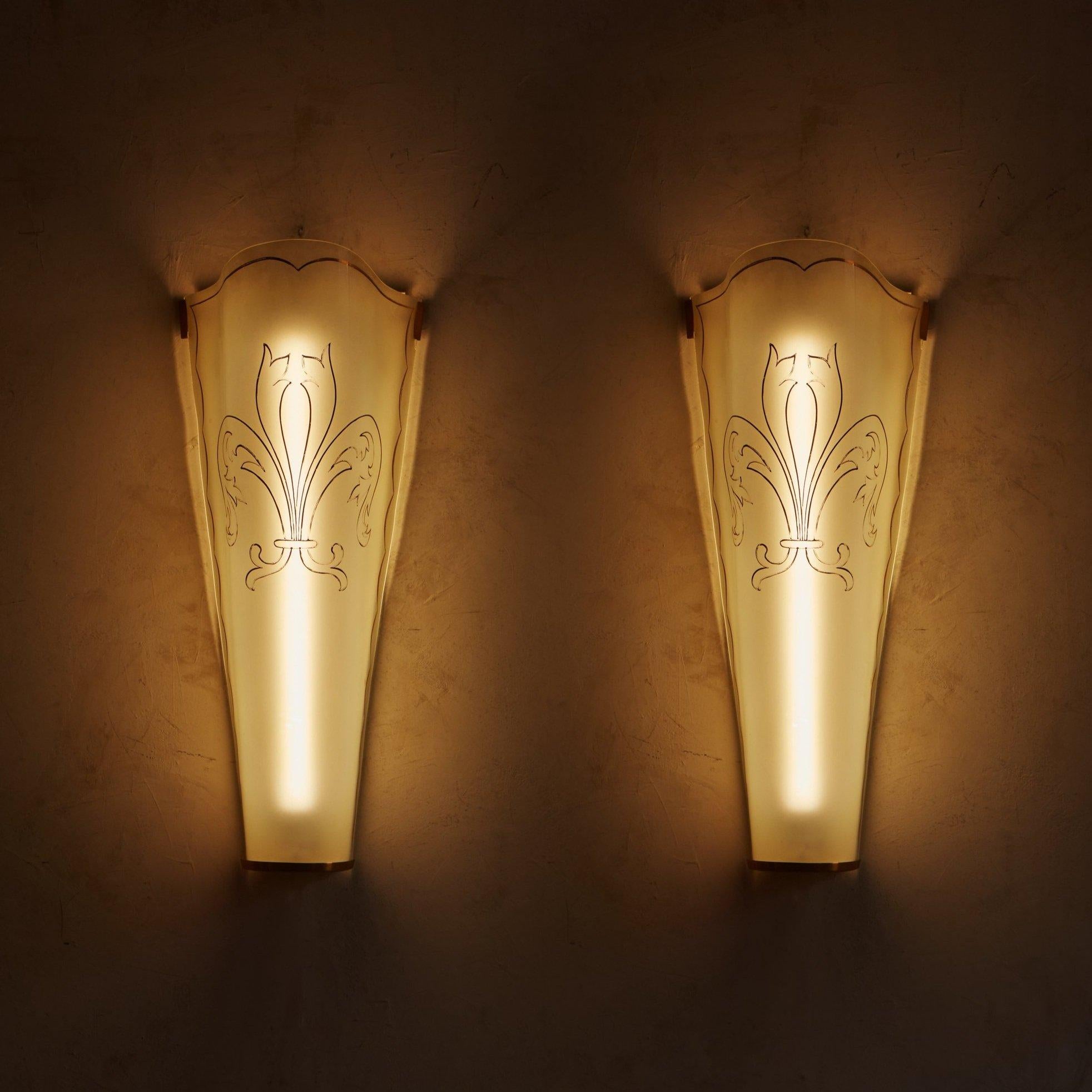A pair of 1930s French sconces featuring tall, curved glass shades with a demilune brass base and hardware. Each shade has an elegant scalloped trim and overlaid gold pattern, which becomes beautifully backlit when these sconces are turned on.