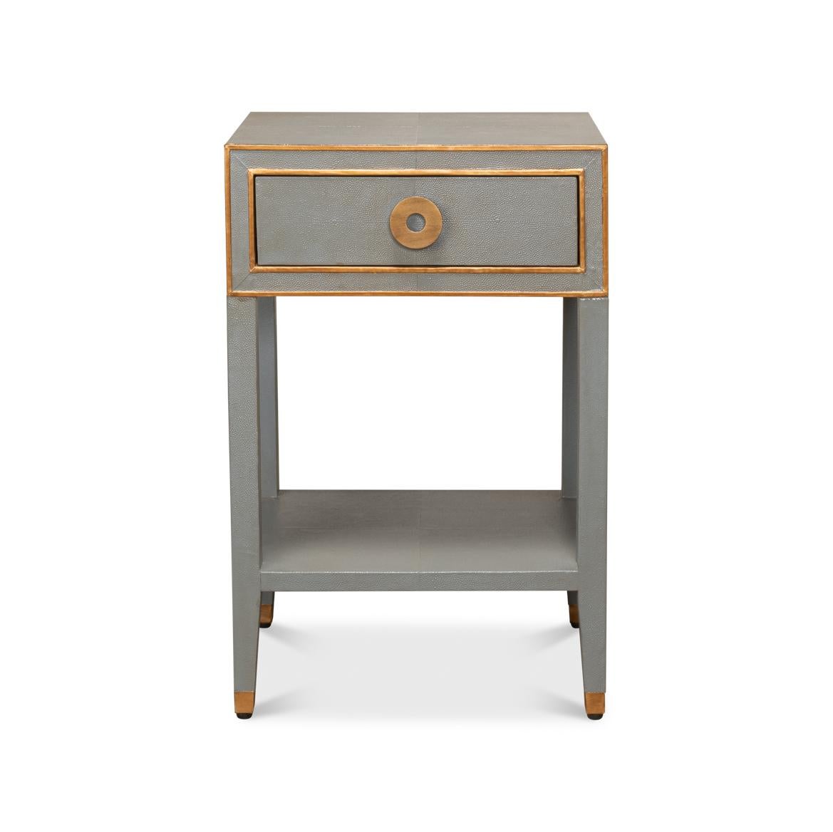 La table de nuit grise associe des surfaces sophistiquées gris tempête à texture de galuchat à des accents dorés pour une déclaration de luxe vintage. La façade détaillée présente des garnitures géométriques en laiton et une tirette circulaire qui