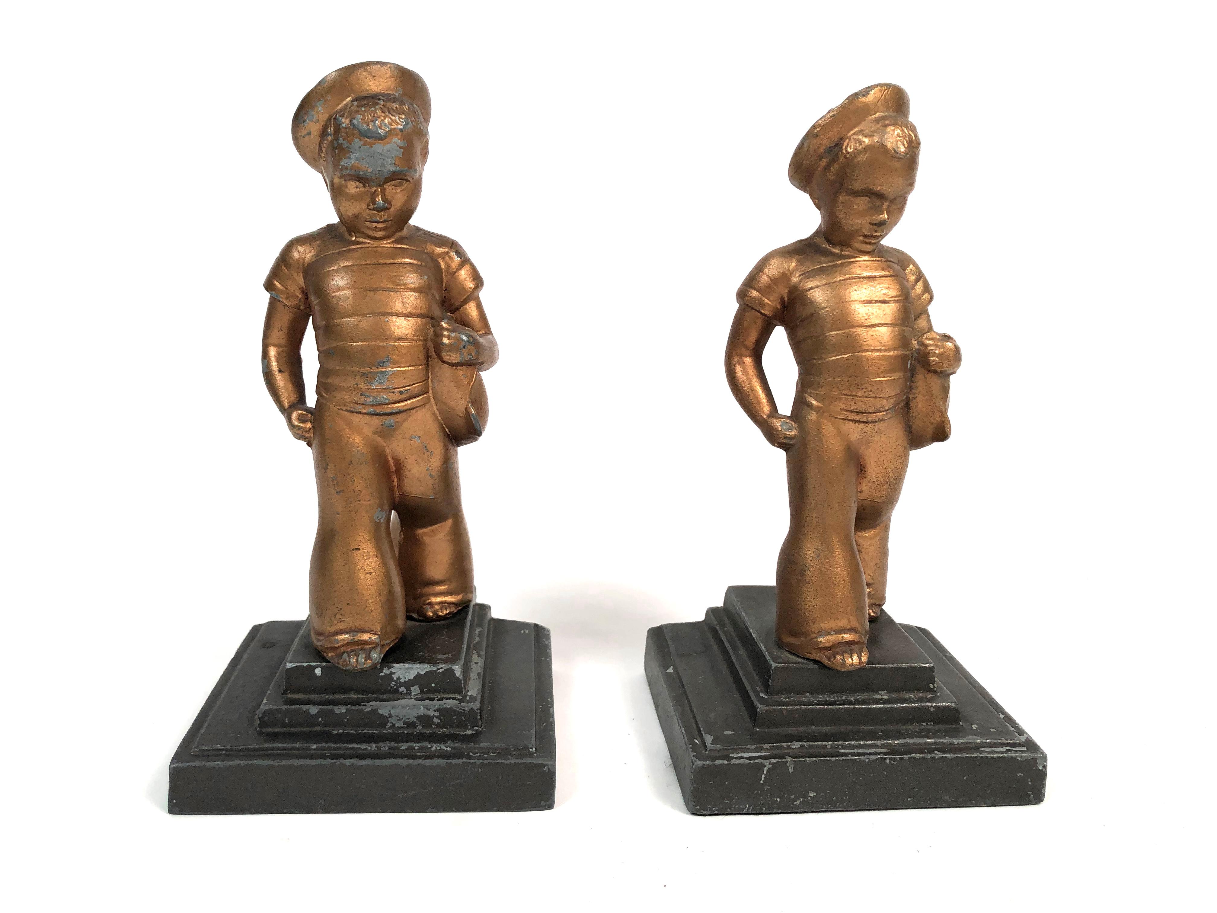 A pair of Art Deco period bookends, each in the form of a charming, stylized little boy wearing a striped sailor shirt, hat and pants, holding a boat under his arm, in gold painted cast metal, on stepped rectangular grey black painted plinths with