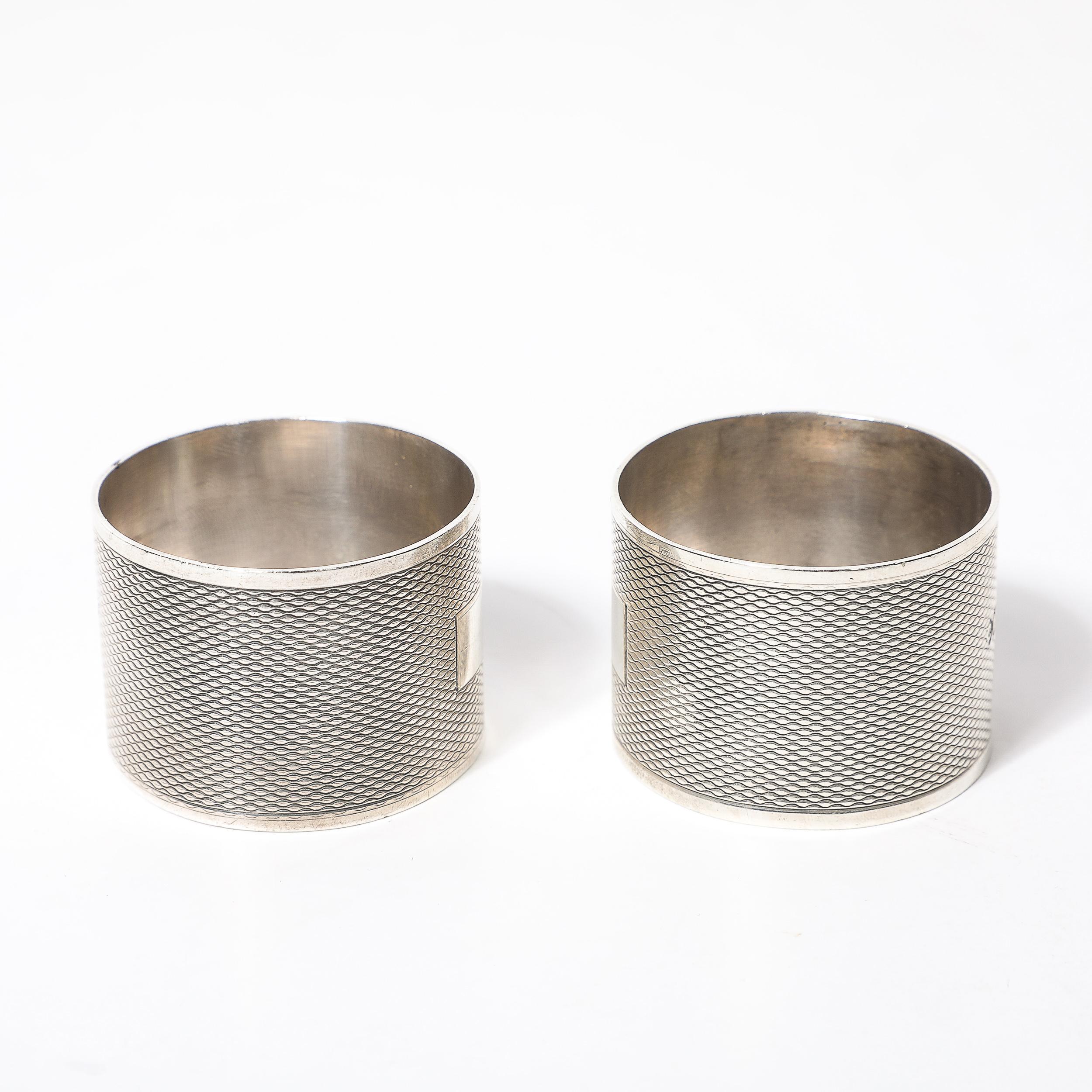 Este elegante par de servilleteros de plata de ley Art decó con detalles de giro de motor, fabricados en Inglaterra hacia 1925, presentan intrincados detalles de textura tejida que añaden un toque distintivo a cualquier servicio de mesa. Su fina