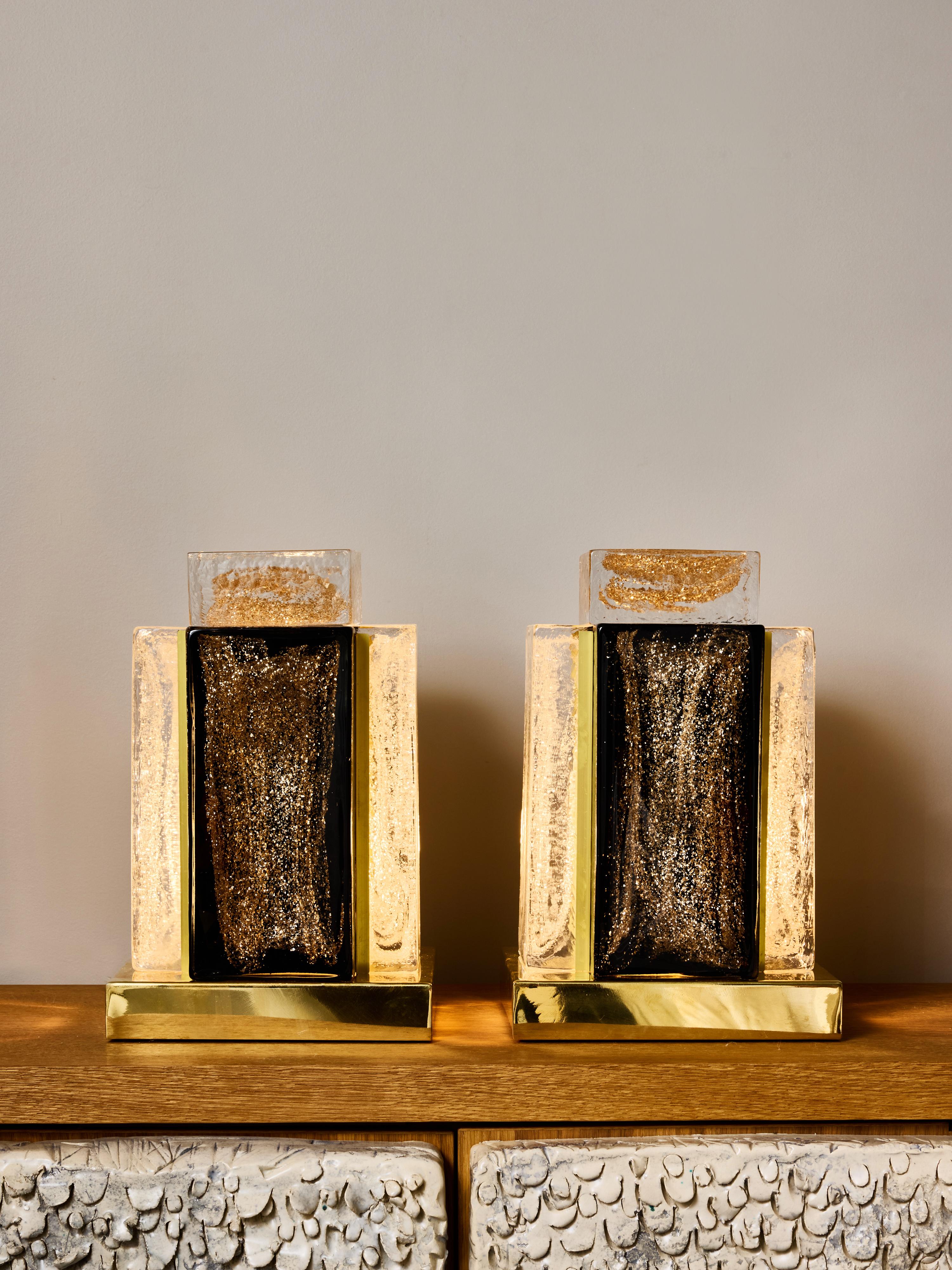 Este par de lámparas de mesa contemporáneas se inspira con elegancia en la geometría clásica del Art Déco. Cada lámpara tiene una base de latón pulido que sostiene cuatro gruesos paneles de cristal dispuestos en rectángulo. Los paneles, dos