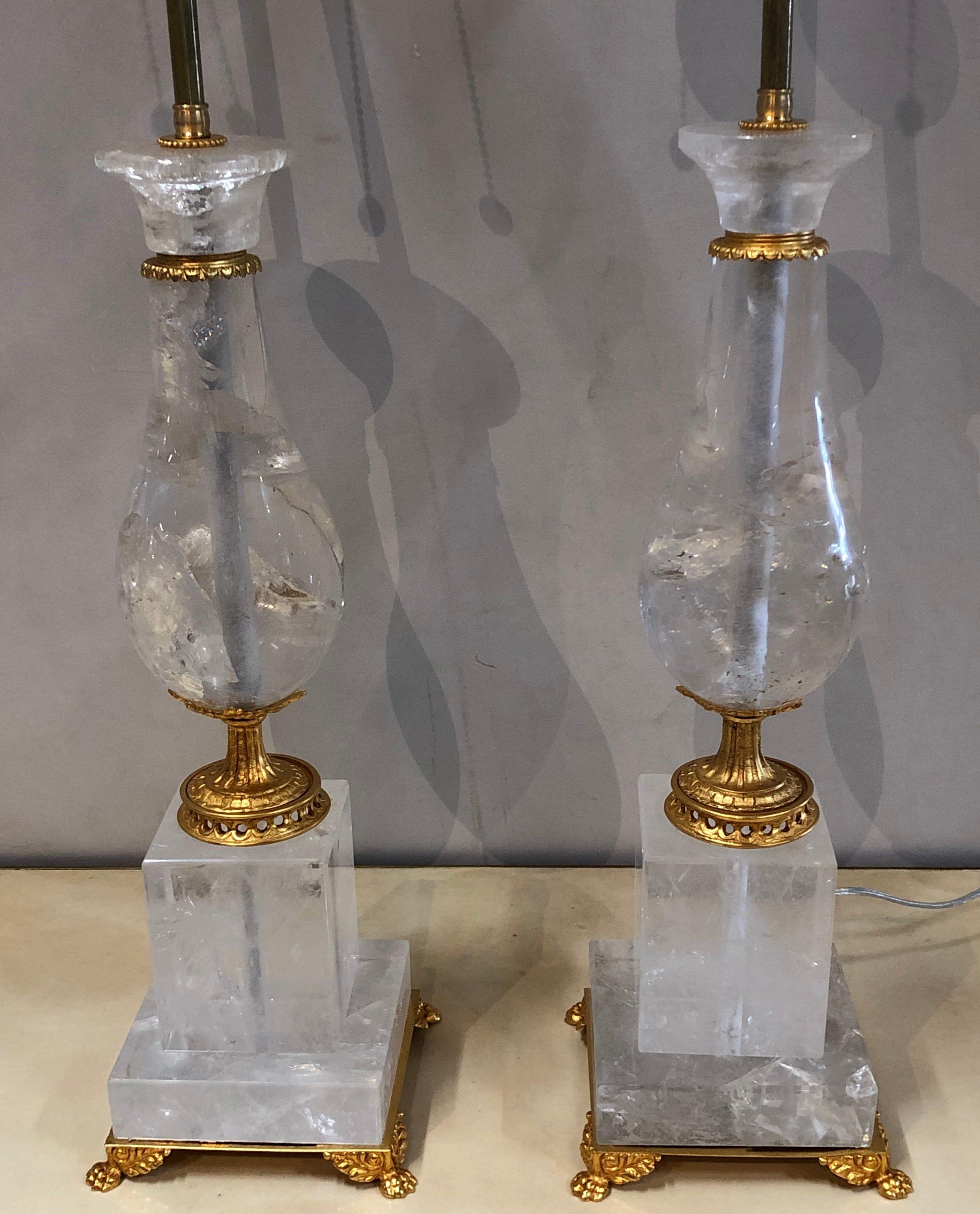 A pair of Art Deco style large rock crystal and brass urn on base form table lamps. The large urn itself measures 19.5 inches high. The gilt metal claw feet with winged sides supporting a column form pedestal base leading to a large column form urn