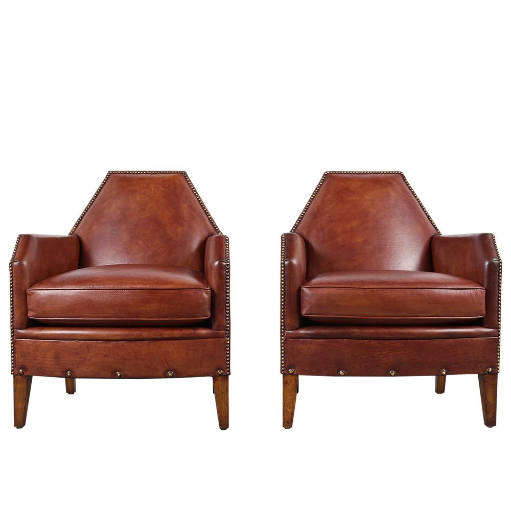 This pair of vintage Art Deco style club chairs features leather upholstery, single cushion, and brass nail details along the edges of the chairs. The chairs has its original leather which is in very good condition and has been dyed in light brown