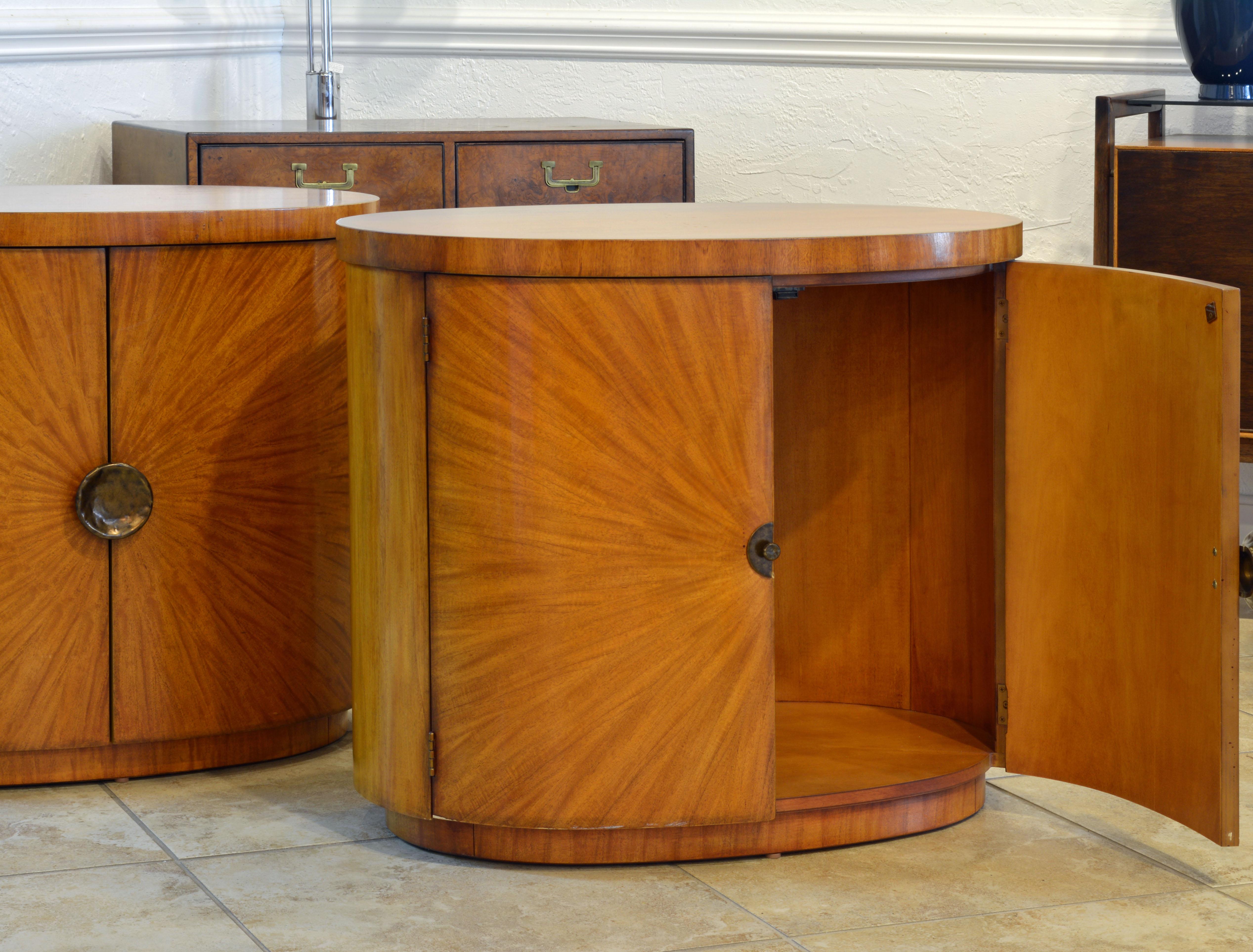 This rare pair of oval Art Deco style light mahogany cabinets or tables are finished all the way around and can be used free standing with great effect. The magnificent doors are sunburst configured and accented by hammered patinated brass handles.