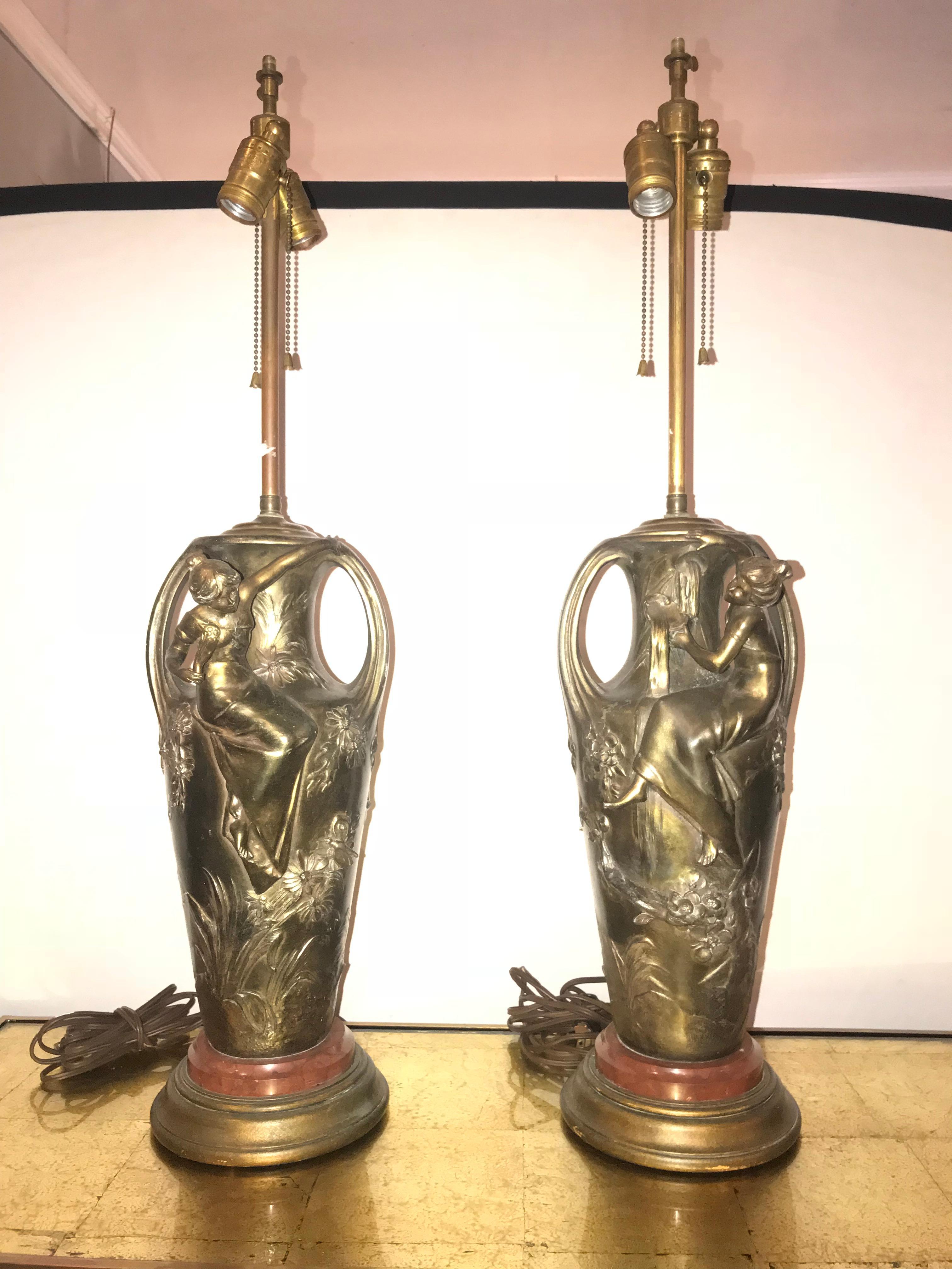 Pair of Art Nouveau figural urns mounted as lamps. These wonderfully cast urns are mounted as table lamps. Each having a young maiden climbing onto a large urn with flowers, vines and the like detailed in a wonderful gold finish.