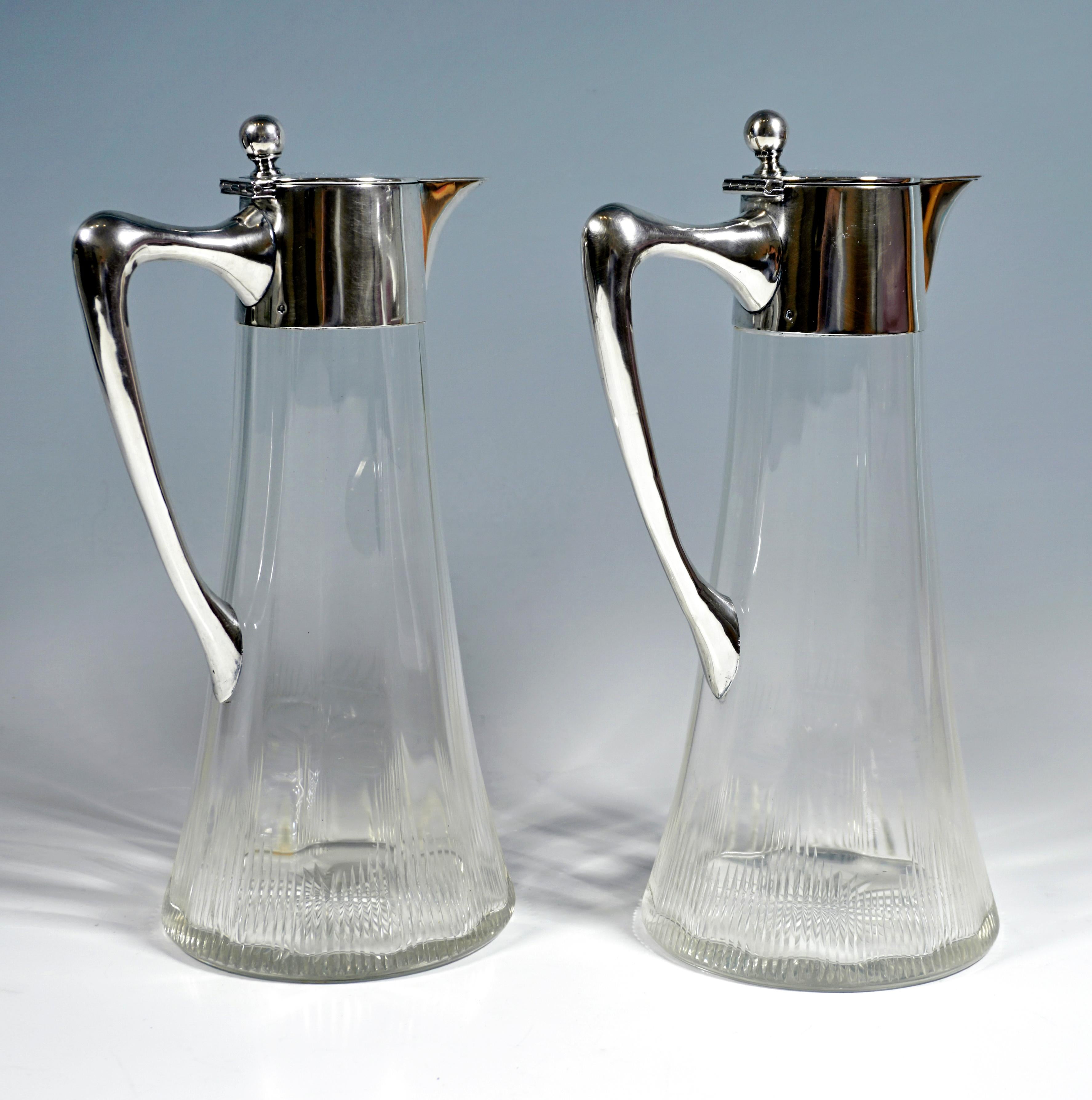Two carafes made of clear glass with a conical body, a twelve-point, wavy thickened wall on the inside, decorated near the bottom with parallel cuts in a serrated shape, a bottom star, smooth silver fitting with a J-shaped, curved handle, covered