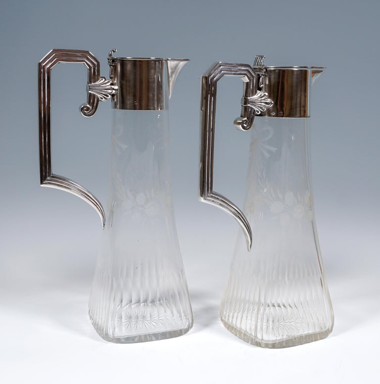 Pair of carafes made of clear glass on a square stand, tapering towards the top to a round cross-section, ground in the lower quarter with an arc-shaped facet, in the upper half delicate flower bouquets and festons in matt cut, ground star, silver
