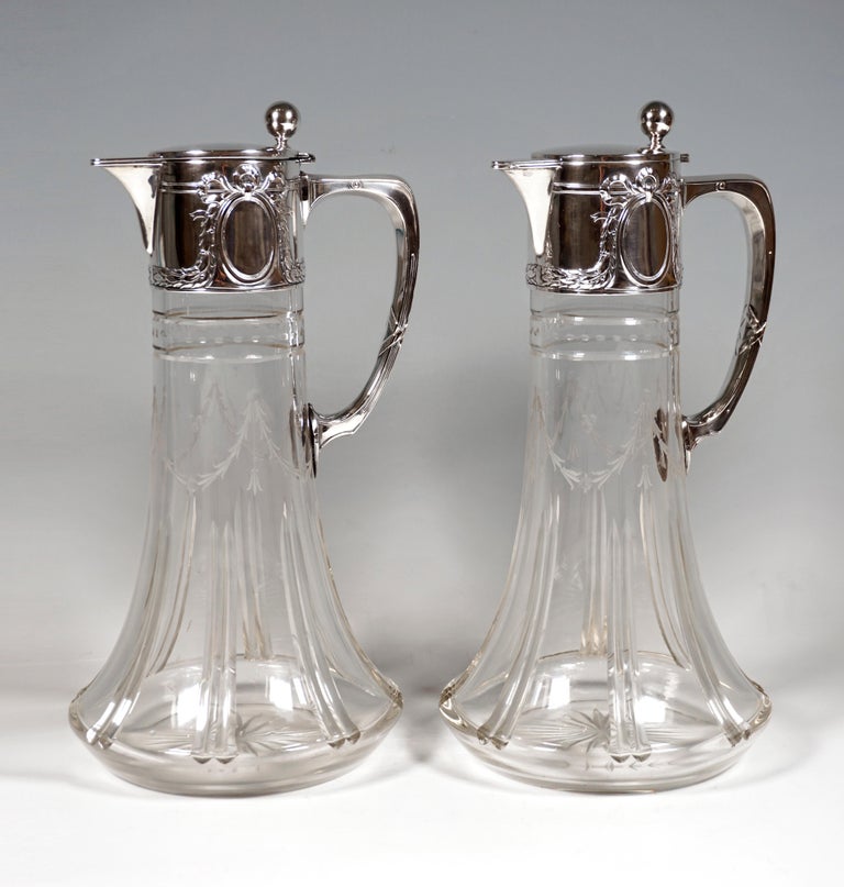 Two carafes made of clear glass with a conical body that swings out at the bottom, pairs of vertically cut lines in the lower half, surrounding leaf garland cut above them, horizontal cut lines and dot decoration on the neck, base star, silver