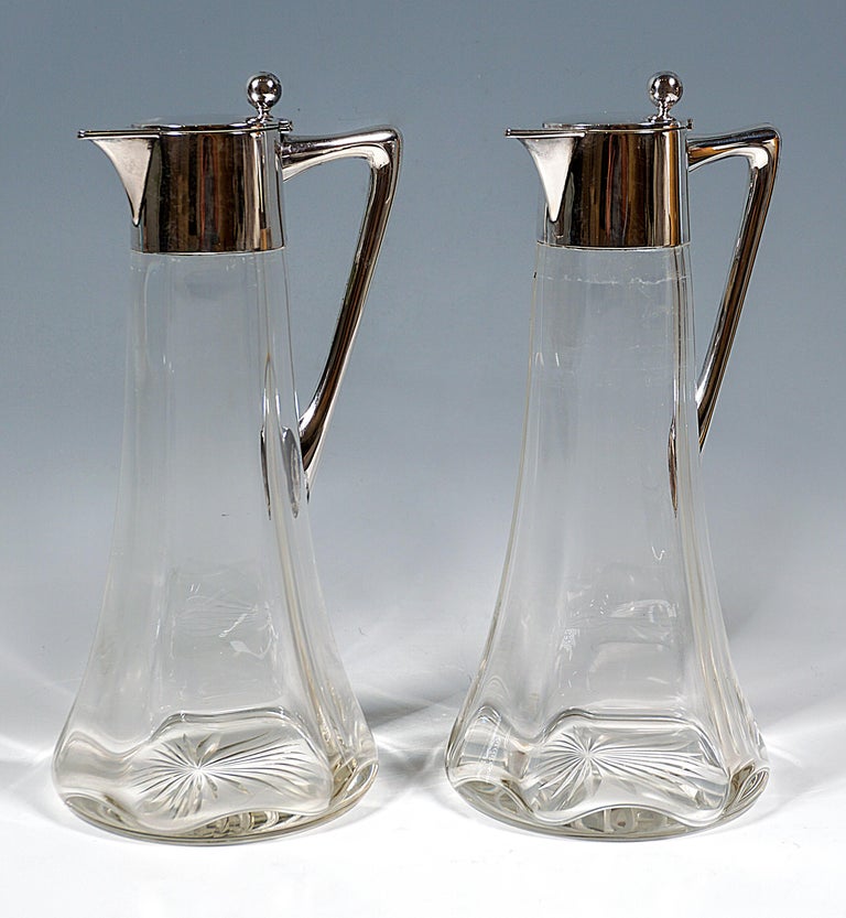 Two carafes made of clear glass with a conical body, inside six-pointed wave-shaped thickened wall, cut base star, smooth silver fittings with J-shaped, curved handle, covered, beak-shaped spout, hinged, straight lid with a ball as a thumb