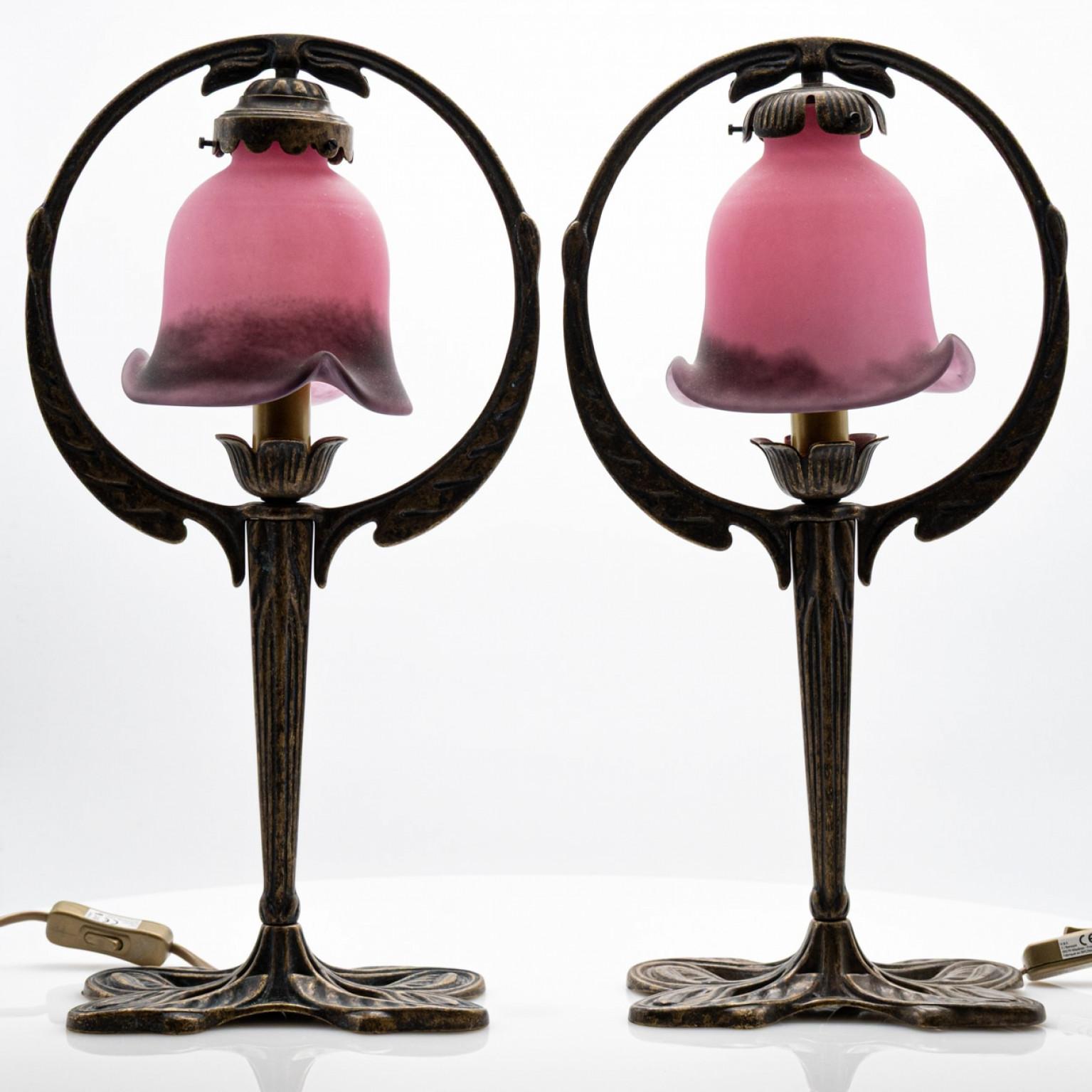 
Si tratta di una coppia di lampade da tavolo in stile Art Nouveau, probabilmente dei primi anni del XX secolo. Il design è caratterizzato da una base in ottone o bronzo con un motivo floreale a forma di giglio. I paralumi sono in vetro rosa o