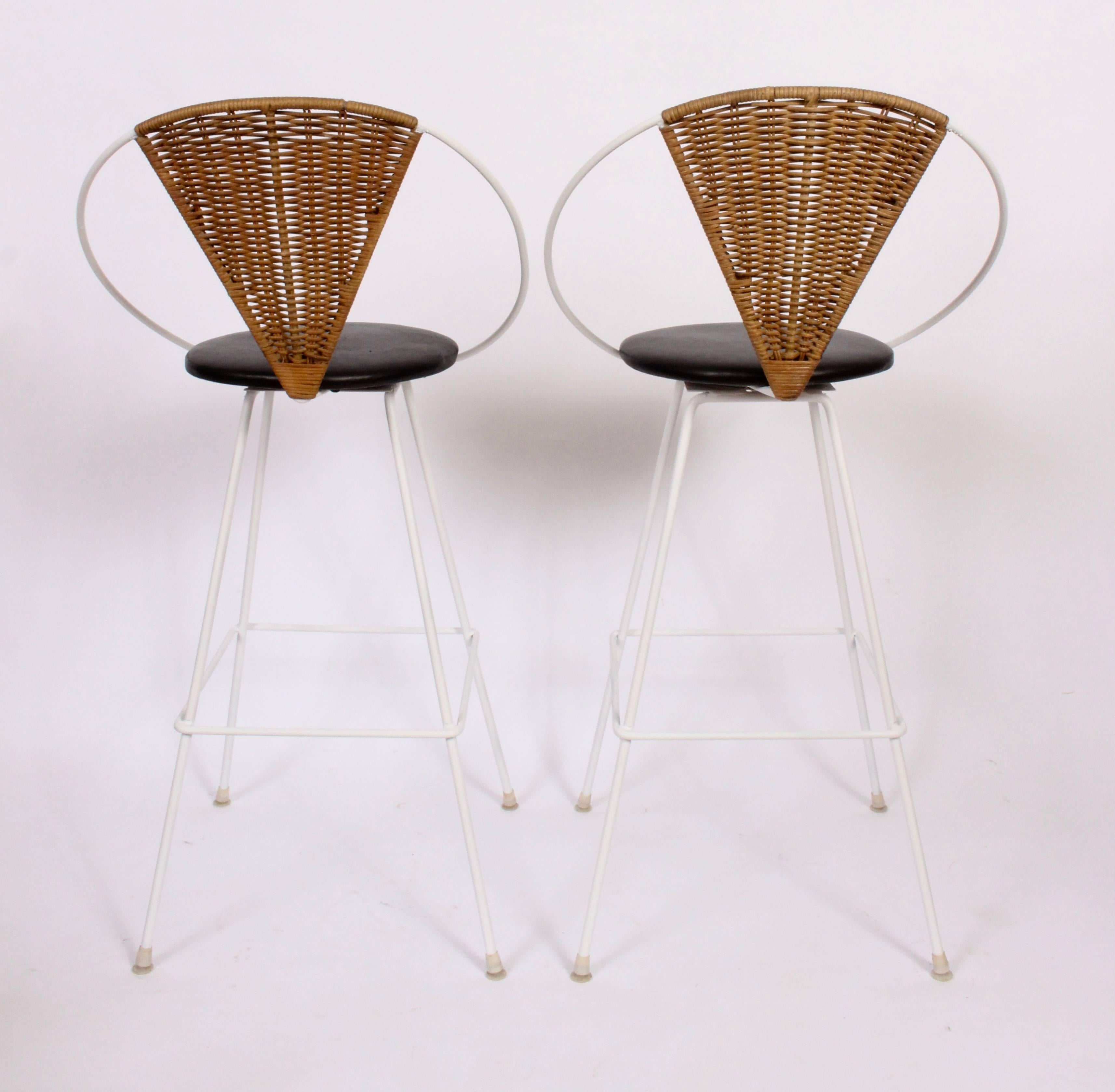 Rare pair of Arthur Umanoff for Shaver Howard Wrought Iron, Rattan Back and Leather Seat Bar Stools. Featuring a White satin enameled wrought iron framework, footrests, hoop back, triangular rattan backrests and newly upholstered round Black leather