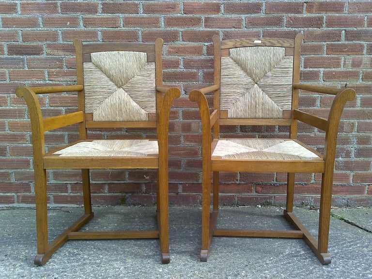 A pair of Arts and Crafts oak elbow chairs, in the manner of George Walton, with re-rushed back and seat panels, square supports with floor stretchers.