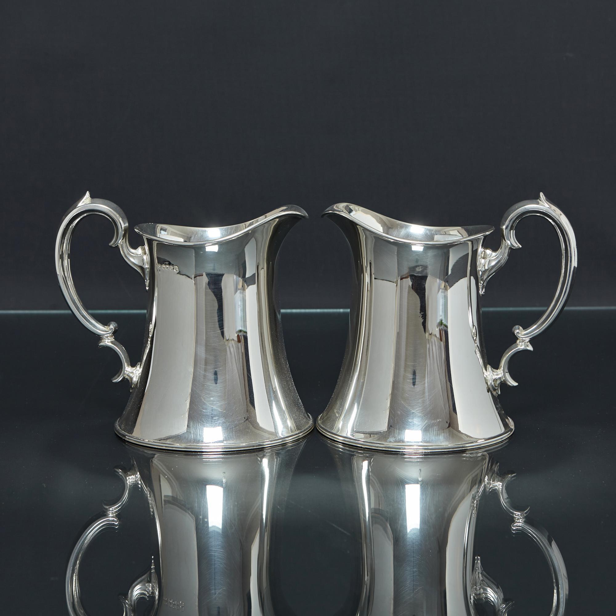 An unusual pair of Arts & Crafts style sterling silver water jugs with waisted bodies and scroll handles. Pairs of water jugs are unusual and this is a particularly stylish example that would complement both traditional and contemporary table