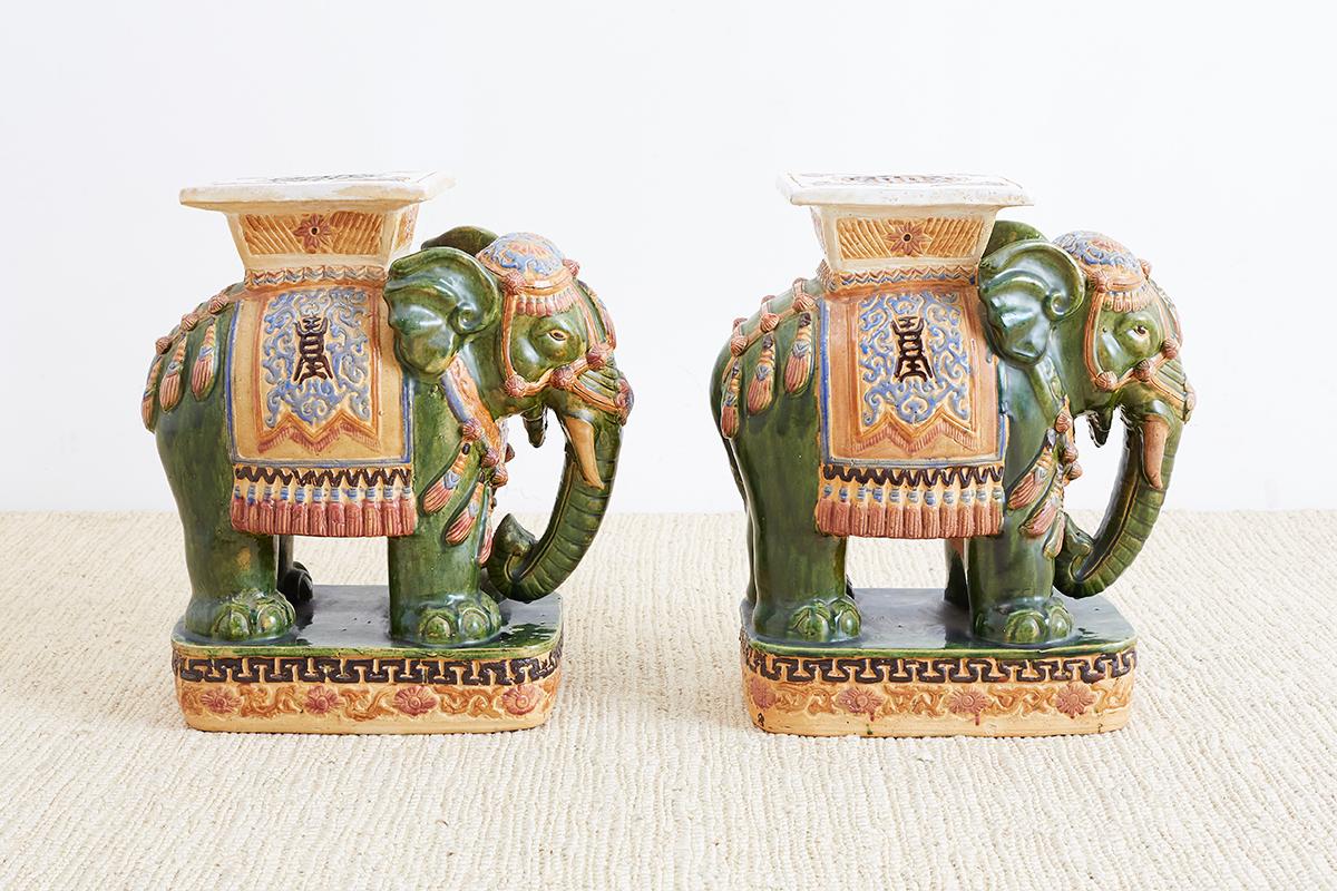 Lucky pair of glazed Asian elephant garden seats or drinks tables. Featuring caparisoned bodies with vibrant colors and good fortune green skin. Each depicted standing on oval plinths with geometric designs.