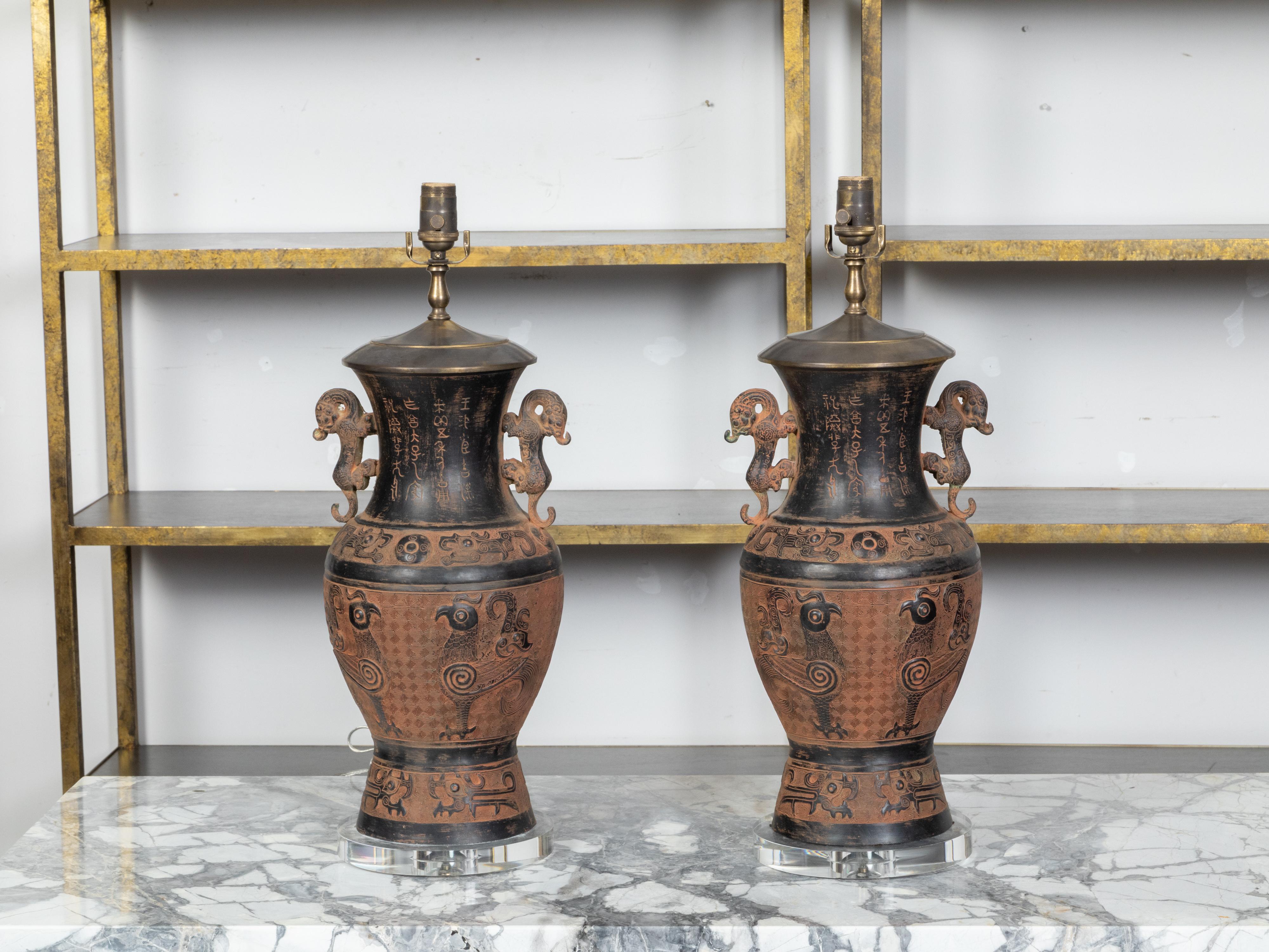 A pair of Asian bronze urn-shaped table lamps from the mid 20th century, with brown and black patina, mythical animals handles, rooster motifs and calligraphy on lucite bases. Created in Asia during the Midcentury period, each of this pair of table