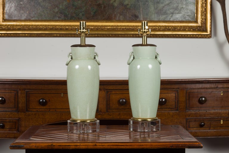 A pair of Asian celadon lamp urns from the mid-20th century mounted on Lucite bases. Created during the midcentury period, each of this pair of table lamps features a celadon urn with slightly crackled finish and lateral ring pulls, mounted on