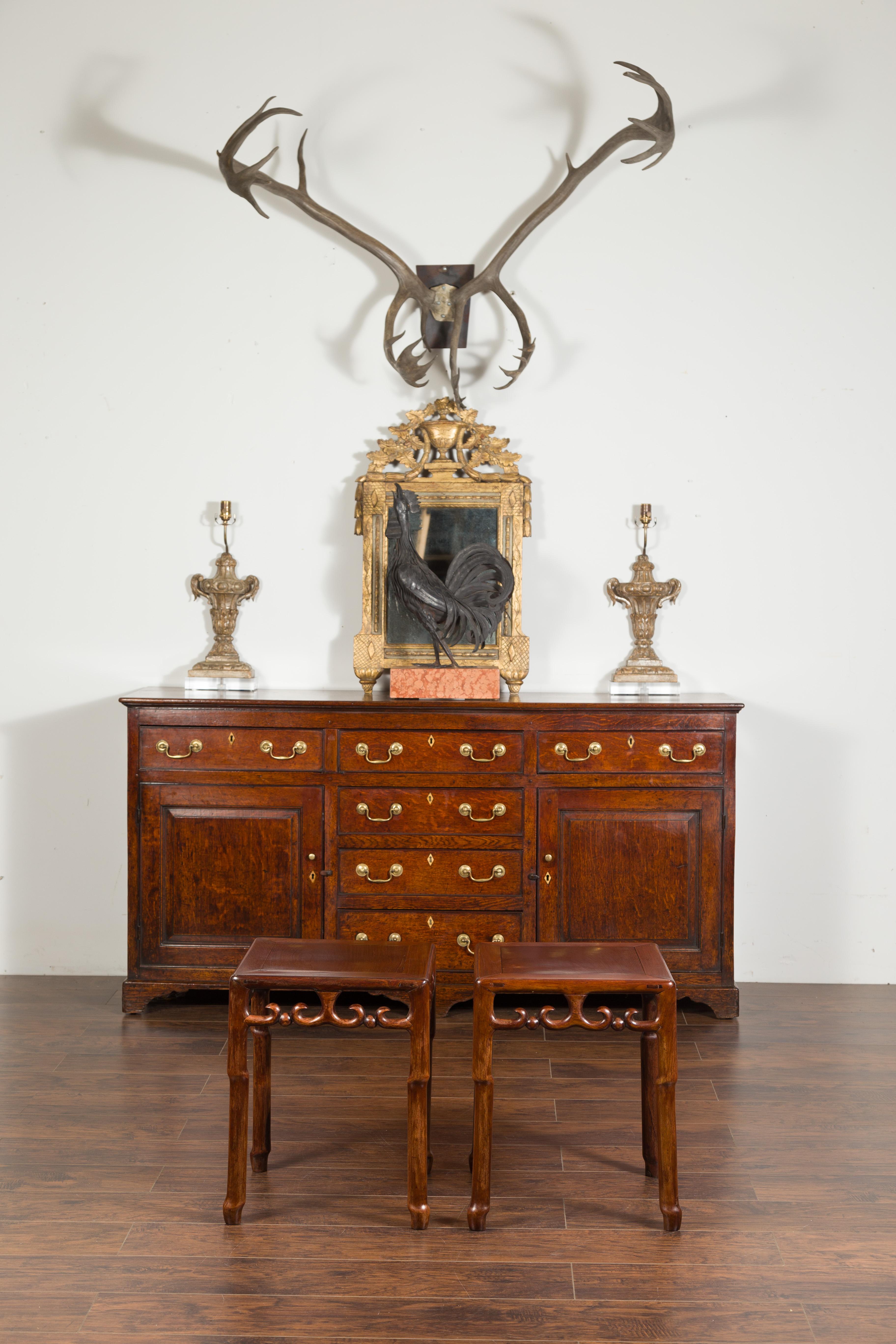 A pair of Asian mahogany side tables from the mid-20th century with scrolling fretwork motifs. Created during the midcentury period, each of this pair of side tables features a square top with central board, resting on an apron pierced with fretwork