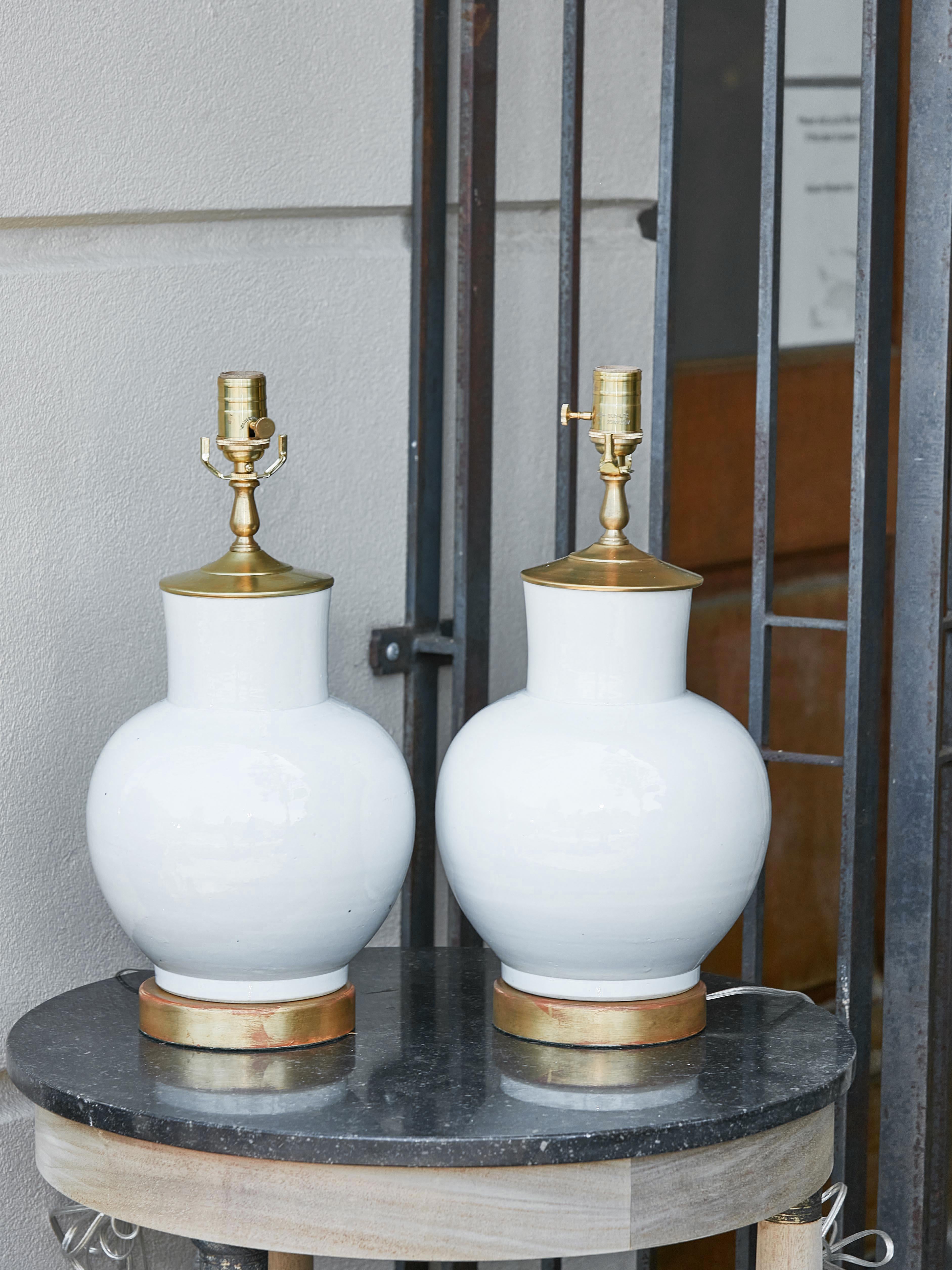 A pair of Asian white porcelain table lamps from the 21st century with brass single lights and circular giltwood bases. Drench your living space in an aura of sophistication and understated elegance with this exquisite pair of Asian white porcelain