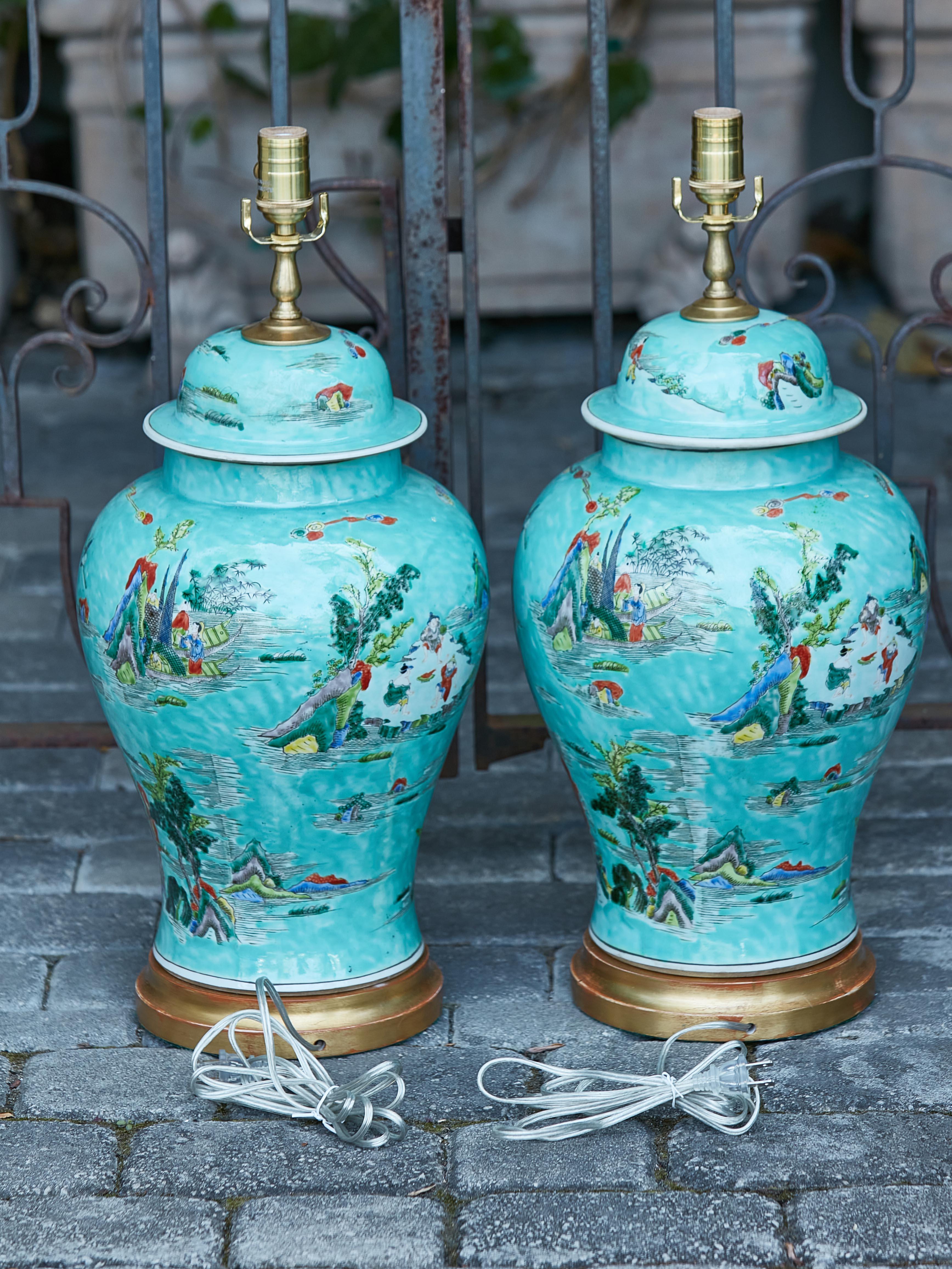 A pair of contemporary Asian lidded urns table lamps from the 21st century with turquoise ground and painted scenes, on circular gilded wooden bases. Infuse your interiors with an aura of Eastern enchantment through this stunning pair of