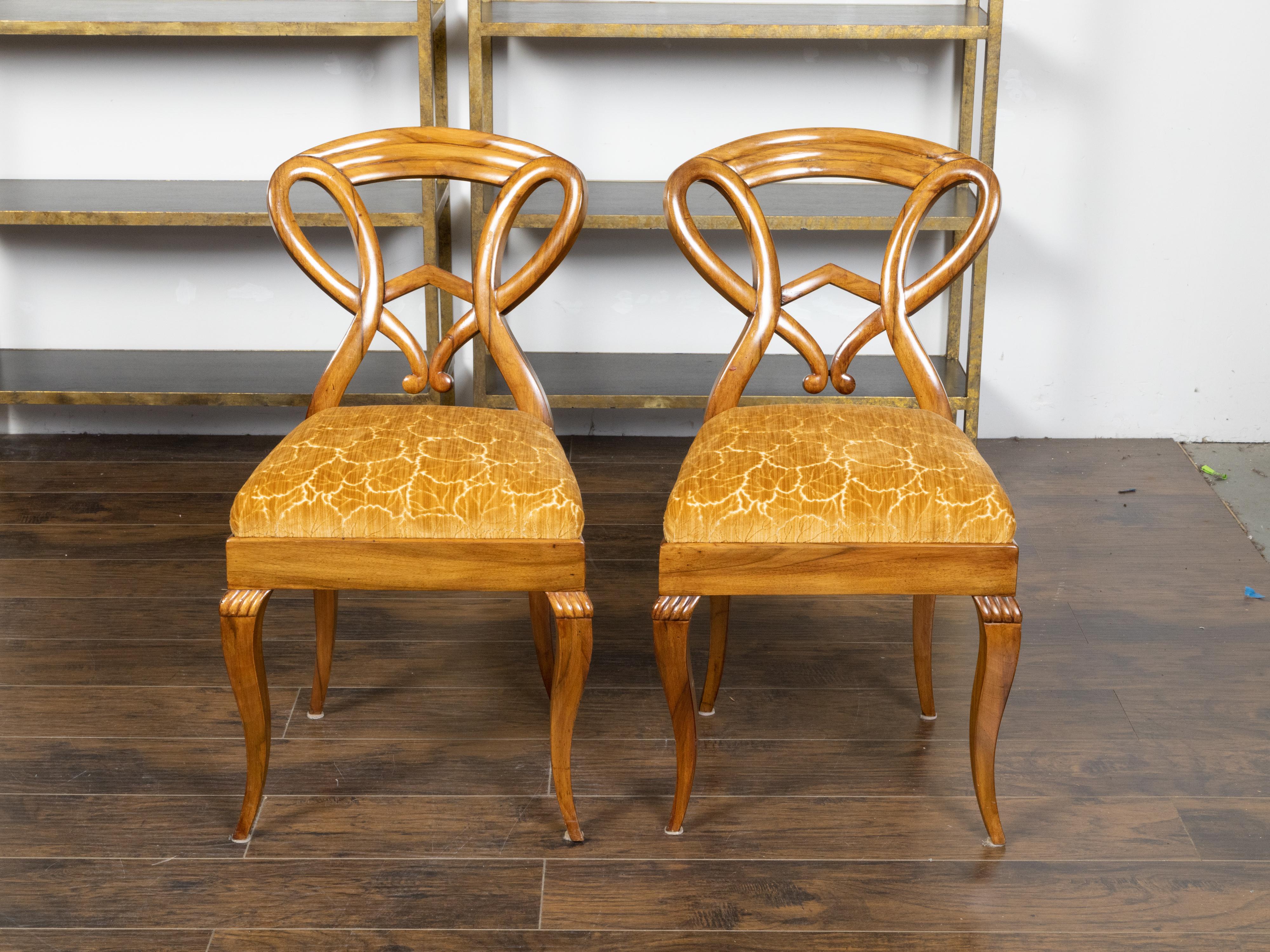 A pair of Austrian Biedermeier period side chairs from the 19th century, with unusual carved open backs, curving legs, carved gadroon motifs and leaf themed upholstery. Created in Austria during the 19th century, this pair of Biedermeier side chairs