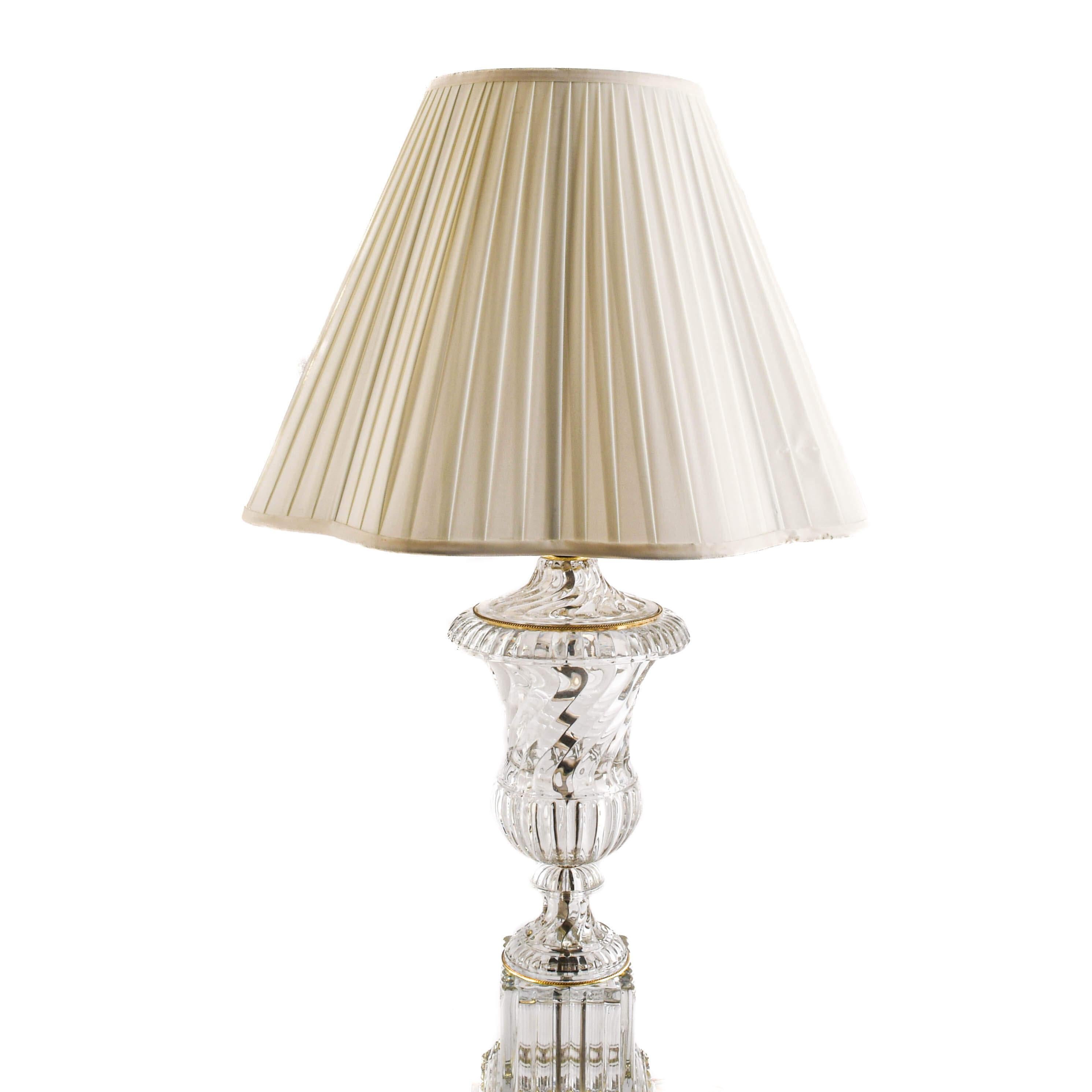 This Pair of Baccarat-Style Crystal Urn Table Lamps on Marble Bases features clear, intricately cut crystal bodies shaped in the form of classical urns, each mounted atop a square fluted glass pillar on a marble base. The crystal elements exhibit