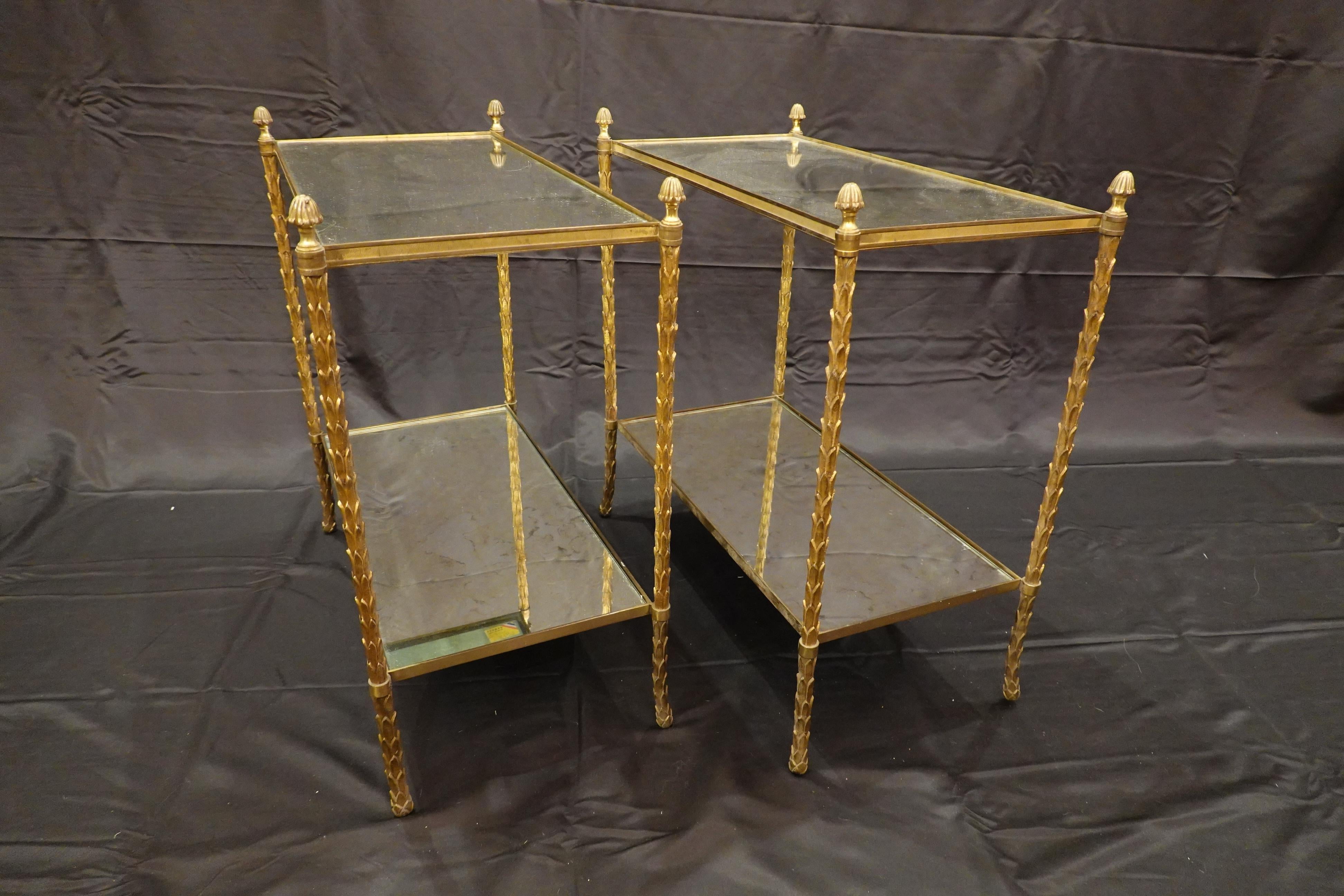 Pair of elegant gilt-bronze two-tiered side tables by French Manufacturer Maison Bagues (circa 1940s-1950s). The legs are in the form of palm fronds terminating in pine cone finials. The eglomise glass tops are slightly smokey with light gold and