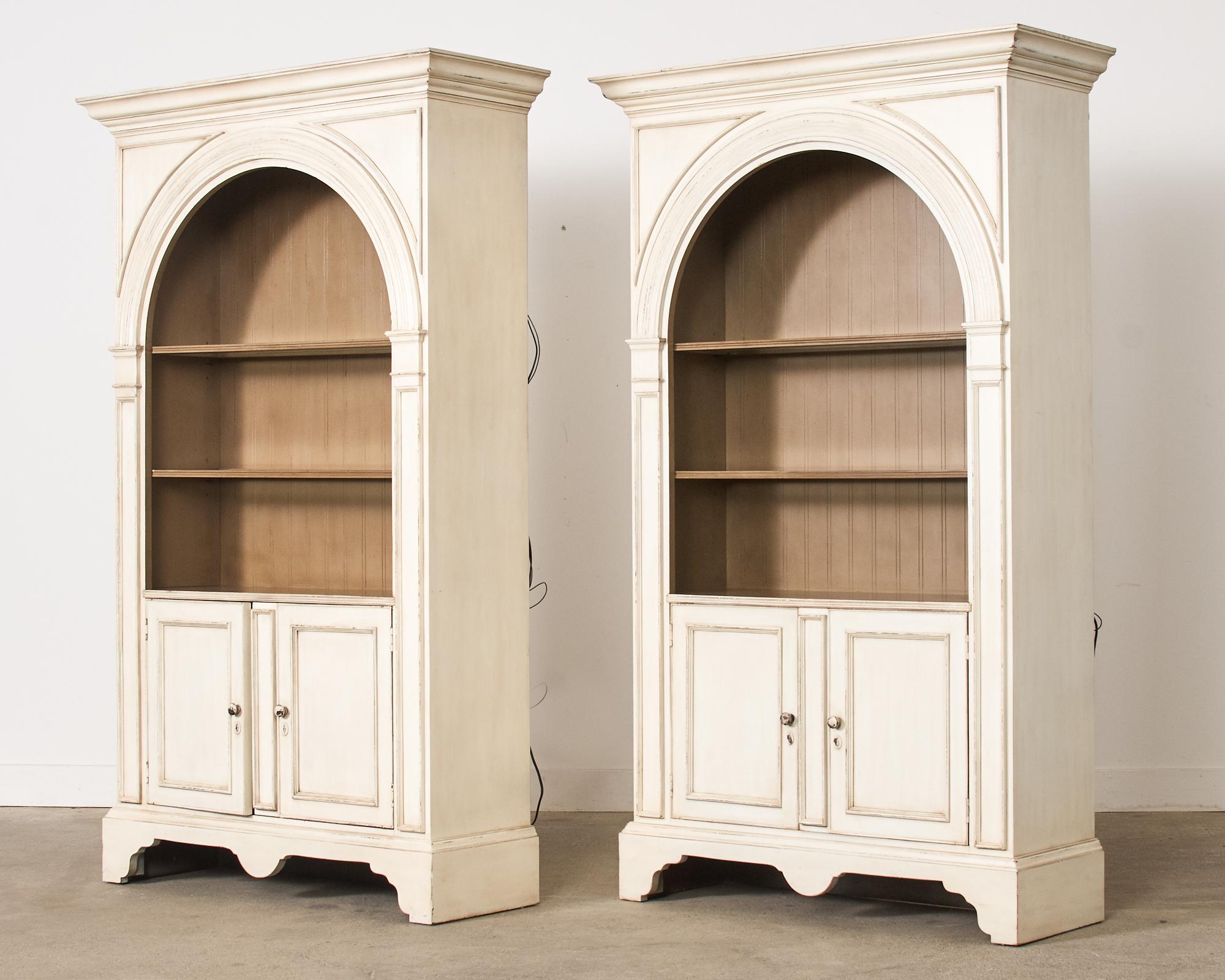 Grand pair of architectural painted library bookcases or display cabinets hand-crafted by Baker. Each large cabinet has an arched opening surmounted by a corniced top. The cases have a fitted three glass shelf interior that is lighted. Below the