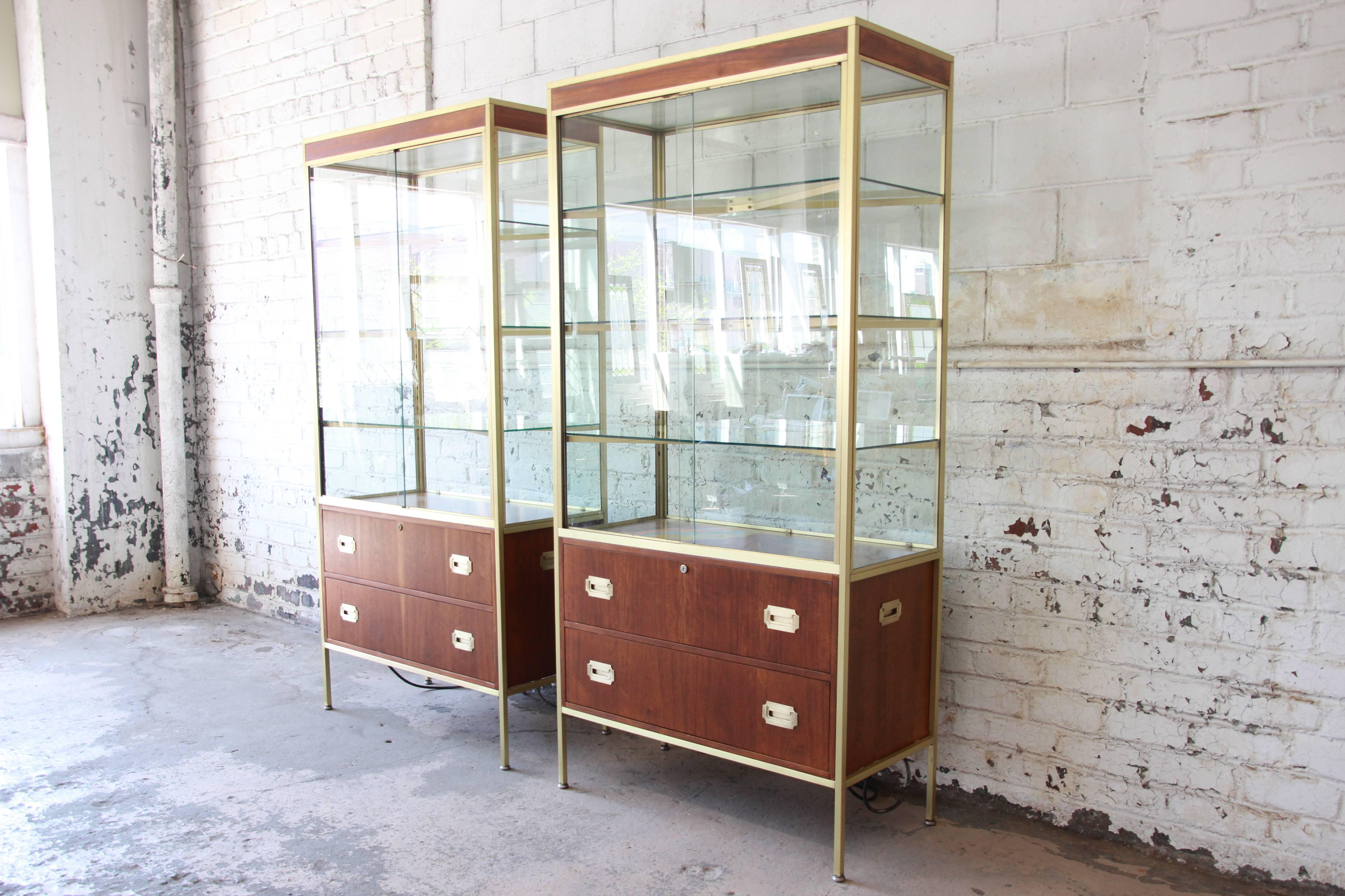 An extremely rare and exceptional pair of midcentury Hollywood Regency Campaign style lighted display cabinets in walnut, brass, and glass by Baker Furniture. The cabinets feature newly polished solid brass frames, with a glass case display and