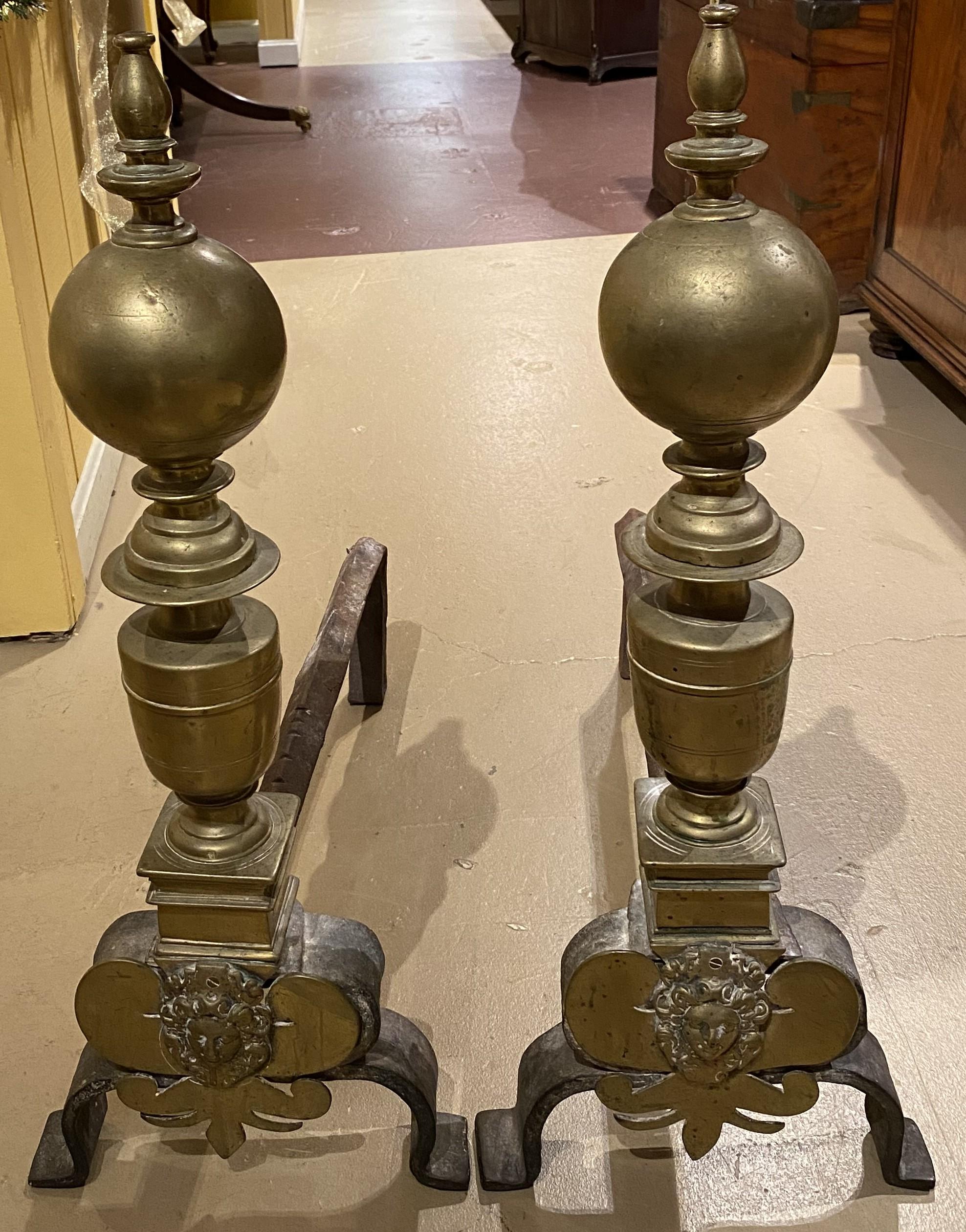A fine pair of heavy Baroque style brass ball andirons with urn form stem and a face decoration on the bases. Probably 19th century Continental in origin, in very good condition, with minor dents, wear and light rust on the dogs, and great overall