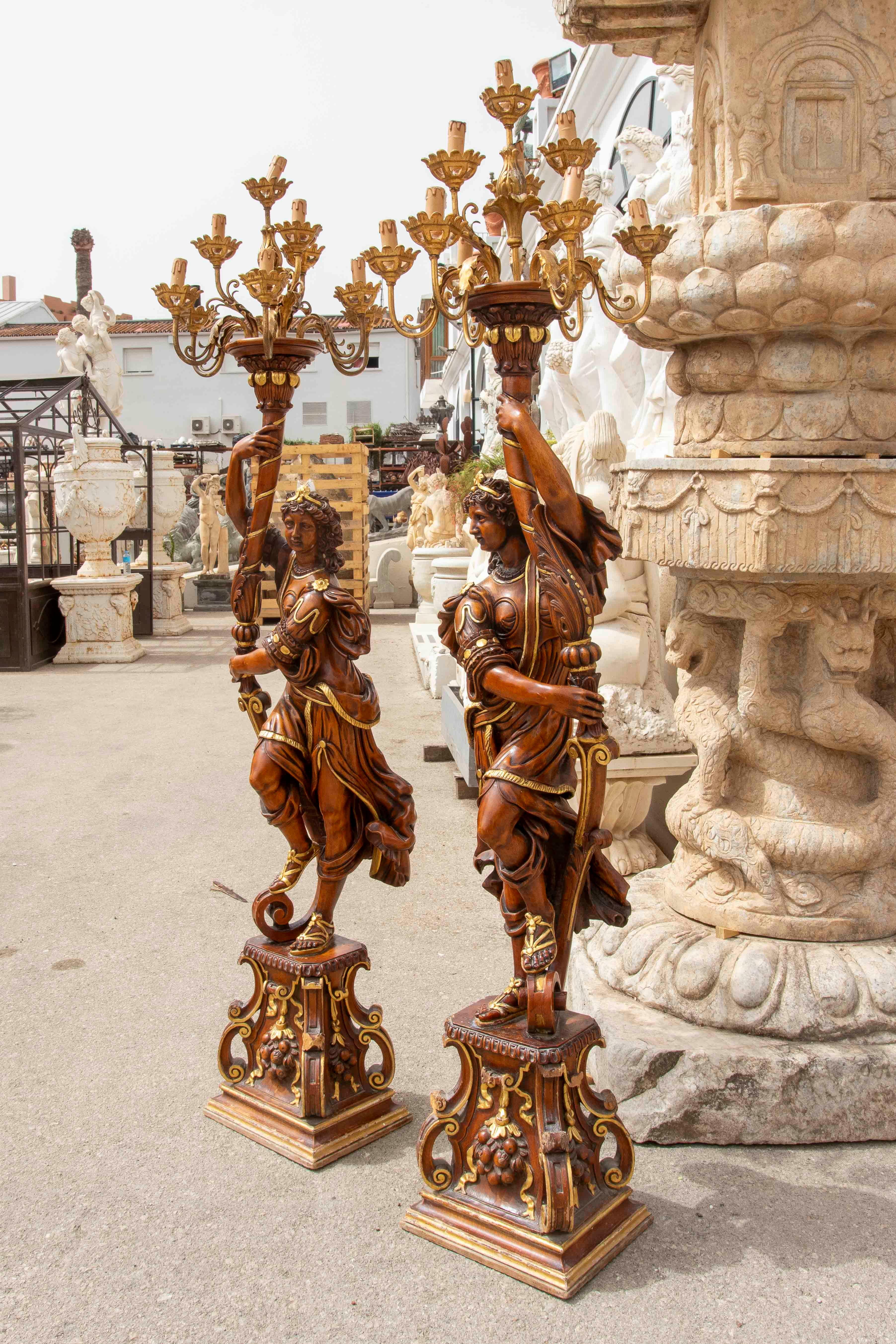 An impressive pair of Baroque candleholders in carved and polychrome wood, depicting allegorical figures in dynamic poses, standing on bases decorated with scrolls and floral motifs. Each figure holds a large, multi-leafed candelabra with gilded