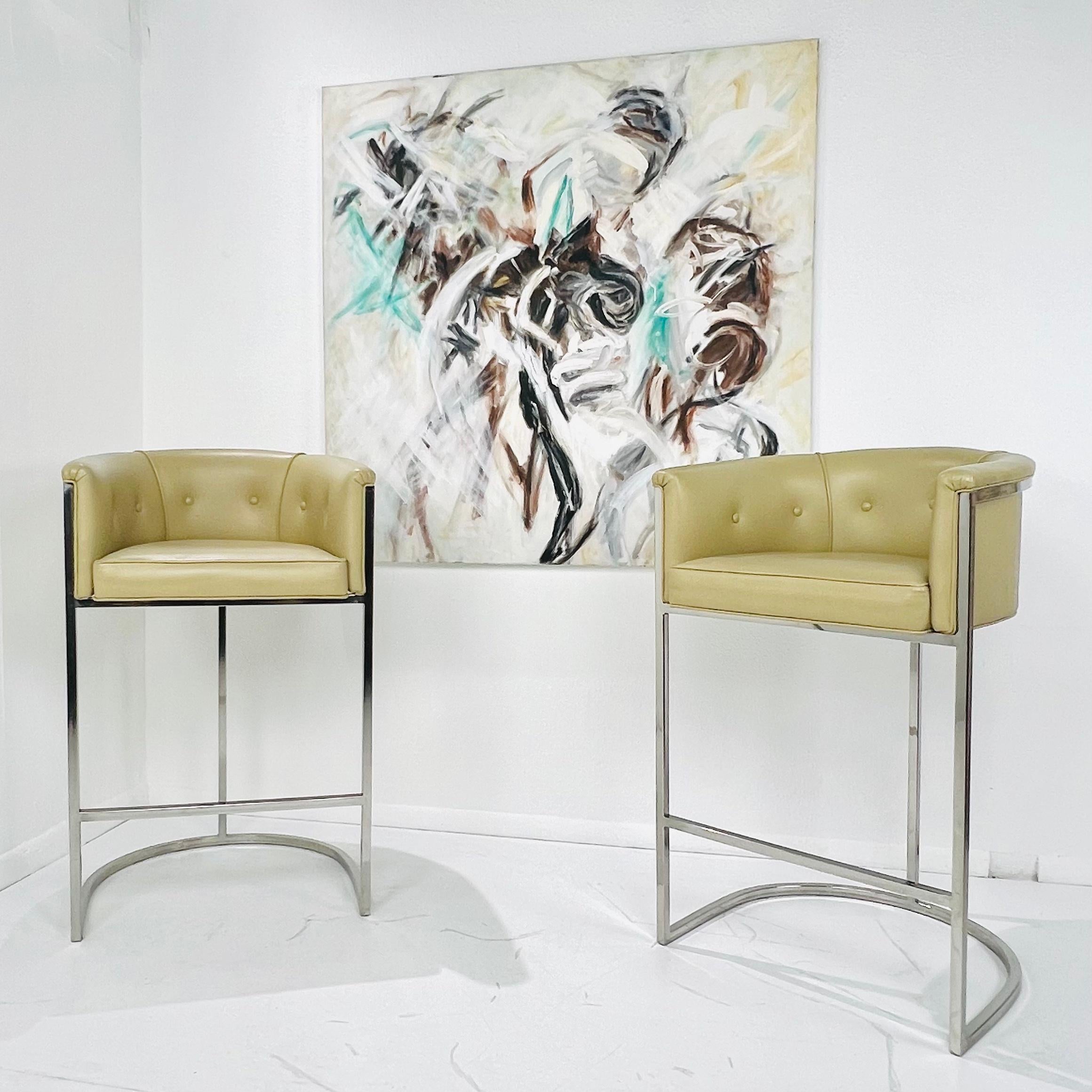 This classic pair of barrel back design bar stools were designed in the 1970s in the style of Milo Baughman. Upholstered in a soft neutral colored leatherette on chrome frames with connecting footrest for maximum comfort. Very good vintage condition