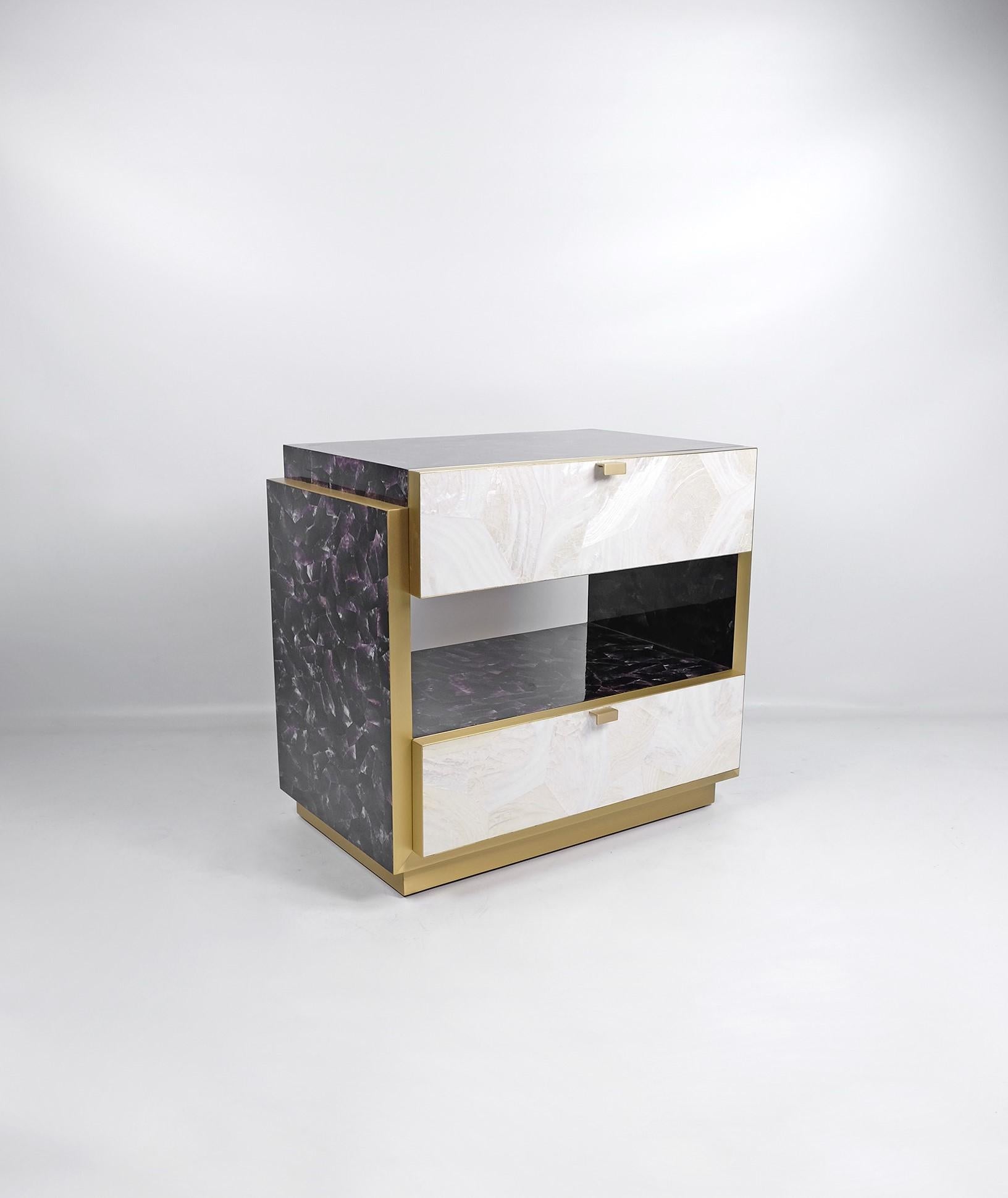 This rectangular bedside table has two drawers and it is made of a purple and white polished shell marquetry.
The edges and the base have brass trims.
The handles are in brass.

The dimensions of this piece are W23.6