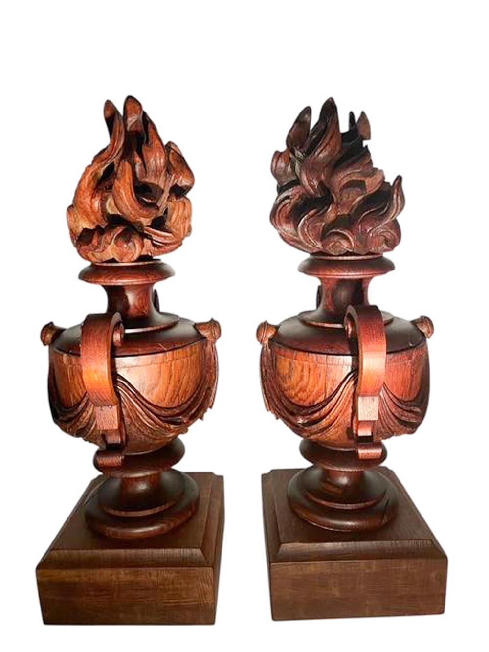 A pair of turned and carved oak architectural elements in the form od swaged urns with handles atop turned socles and set on square oak plinths with molded top edges. Each urn topped with a carved flame finial. the back of each finial is cut away,