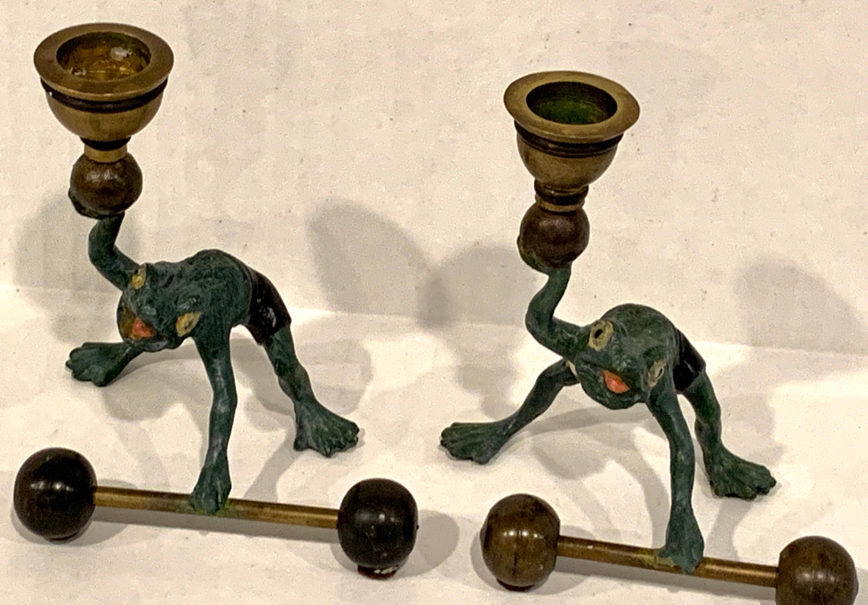 Pair of Bergman style Austrian old painted bronze weightlifting frogs, each one realistically cast and painted, dressed in black and white shorts, balancing a raised shot put and barbell. Each one holds a one standard based candle.