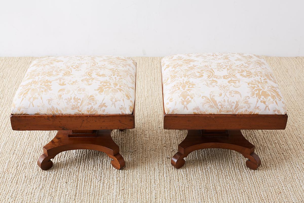 Remarkable pair of hand-carved footstools made in the Biedermeier taste of 19th century Europe. Featuring a Fortuny upholstered cushion in a square case. Constructed with exposed dovetail joinery on the corners and supported by a square column