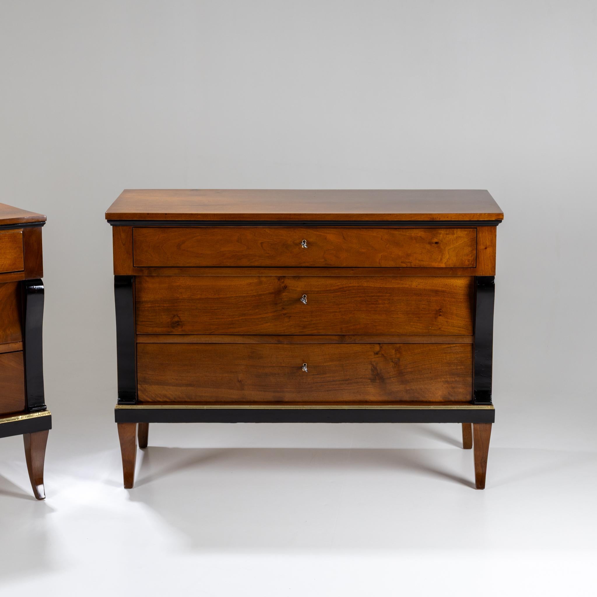 Pareja de cómodas Biedermeier de tres cajones, elevadas sobre patas ahusadas y ligeramente curvadas. Los bordes y las pilastras de voluta laterales están ebonizados. Una discreta banda de latón enmarca la parte inferior de cada cómoda. Hechas de