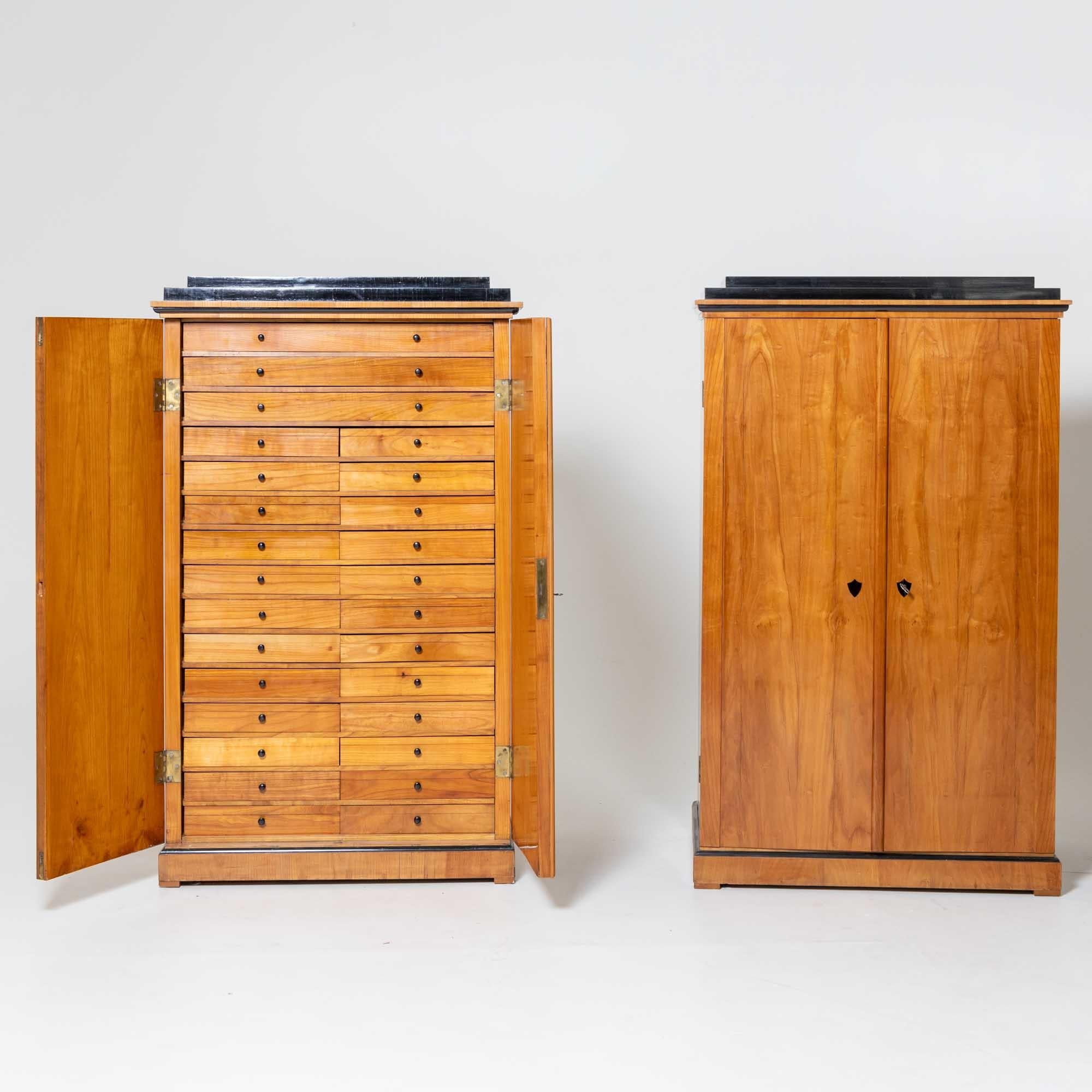 Pair of Biedermeier collection cabinets in cherry veneer with ebonised top plate. The tall two-door cabinets are equipped with a large number of drawers. The drawers are removable and with a glass shelf and glass lid they are ideal for storing even