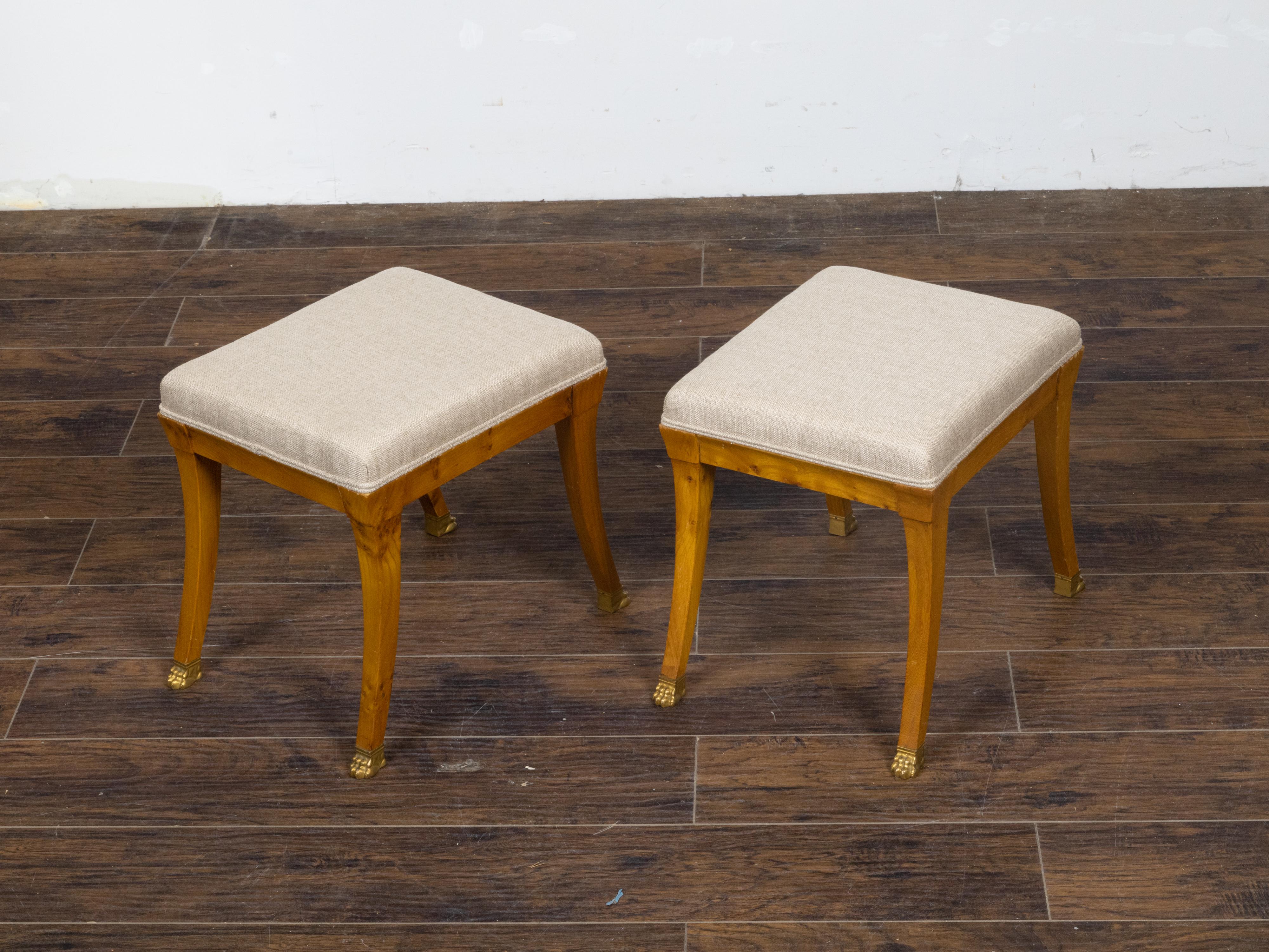 A pair of Austrian Biedermeier period walnut stools from the 19th century, with saber legs, carved giltwood lion paw feet and new custom linen upholstery. This pair of Austrian Biedermeier period walnut stools from the 19th century exudes an