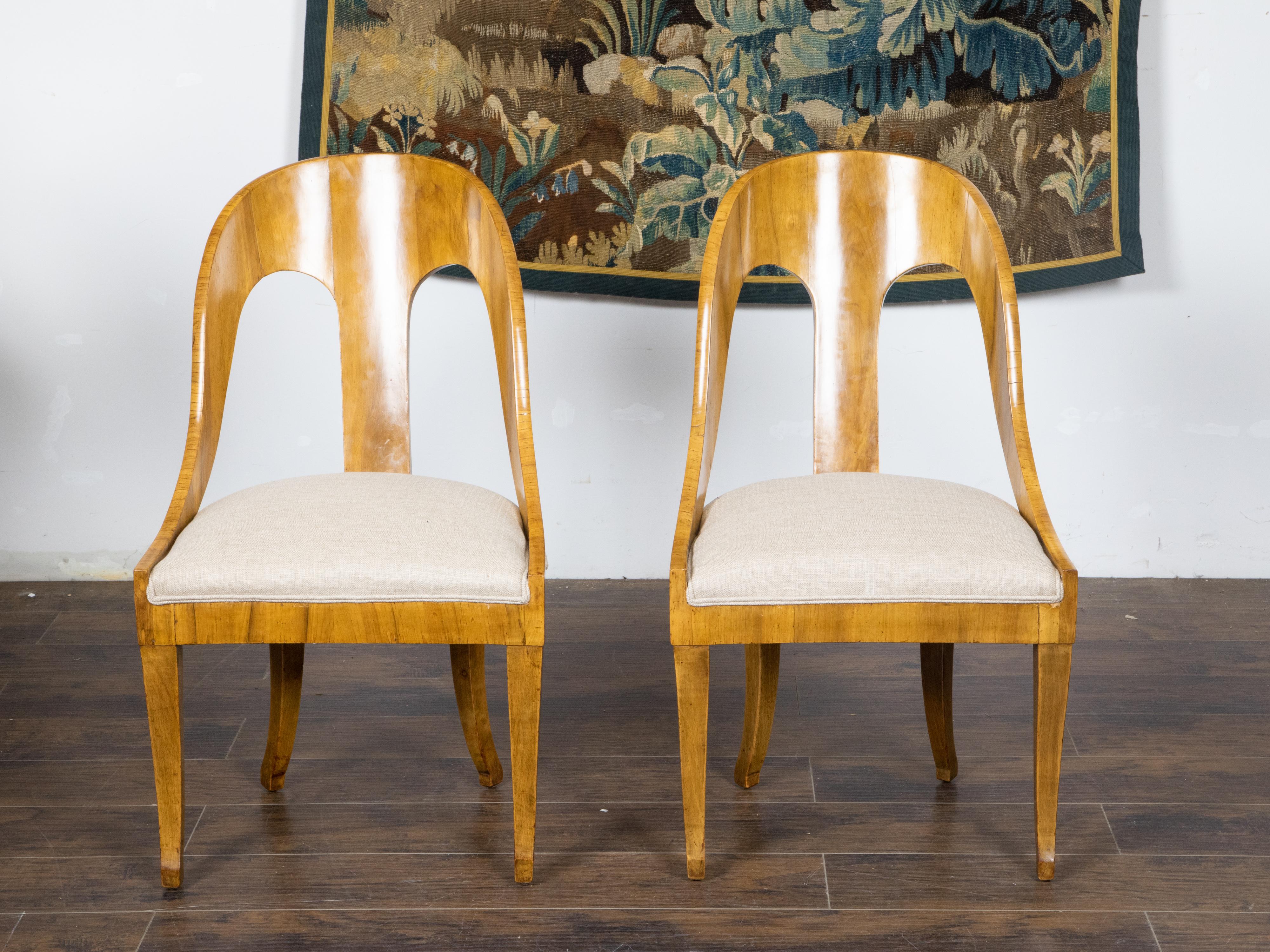A pair of Biedermeier style walnut spoon back chairs from the early 20th century with curving backs, saber legs and new linen upholstery. Created during the Turn of the Century, this pair of walnut spoon back chairs showcases the stylistic