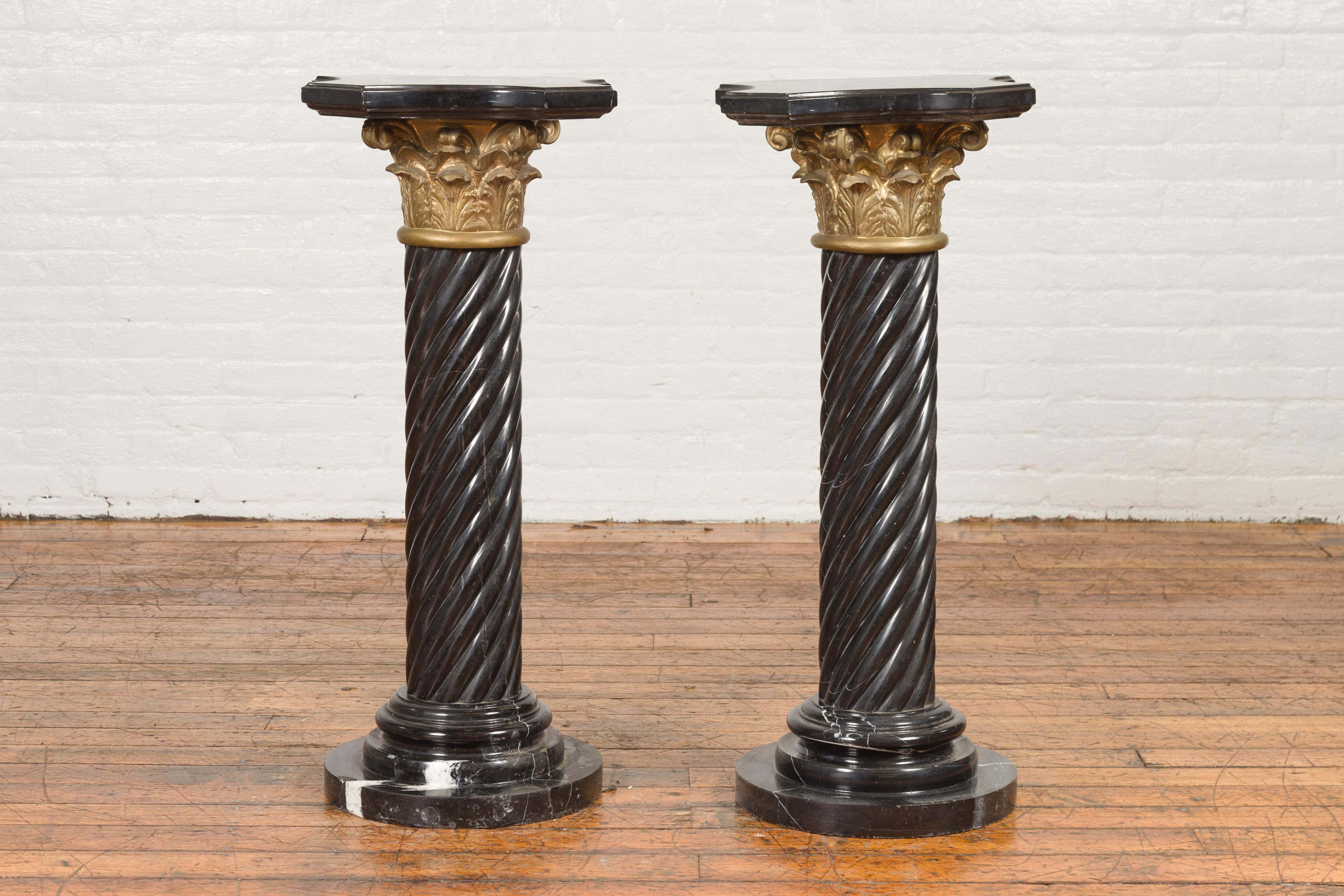 A pair of vintage Thai black Carrara marble pedestals with bronze Corinthian capitals and twisted columns. Created during the midcentury period, each of this pair of pedestals features a shaped marble top with in-curving effects, sitting above a
