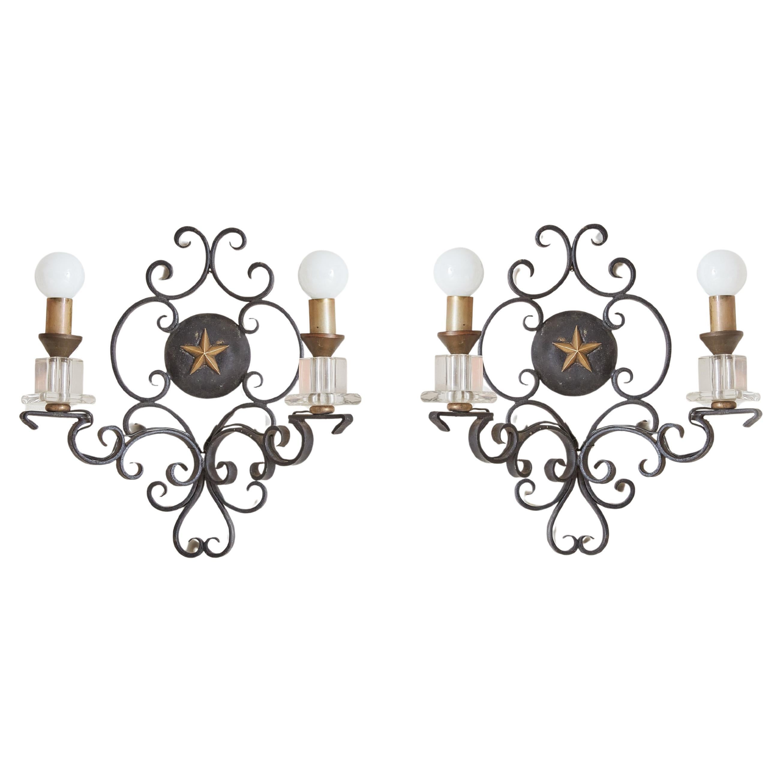 An elegant 1950s French black iron wall sconce featuring intricate scrollwork and a beautiful natural, aged patina. The two-arm silhouette is adorned with brass candle covers, a central star emblem, and stacked geometric glass details. France,